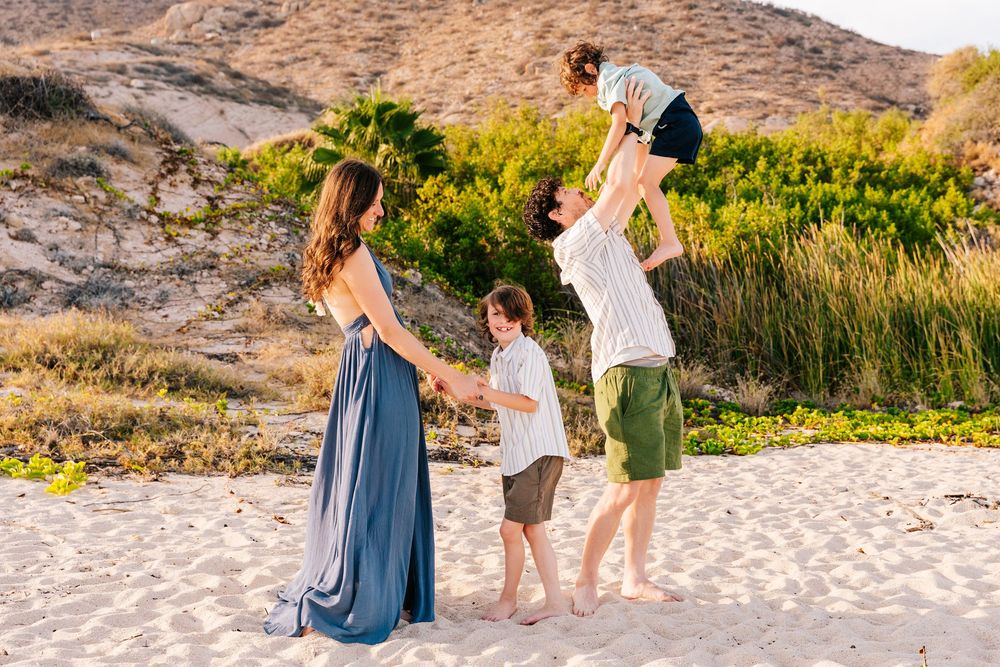 Family of four enjoying a sunny beach day, with dad lifting child overhead while mom and son hold hands on sandy shore.