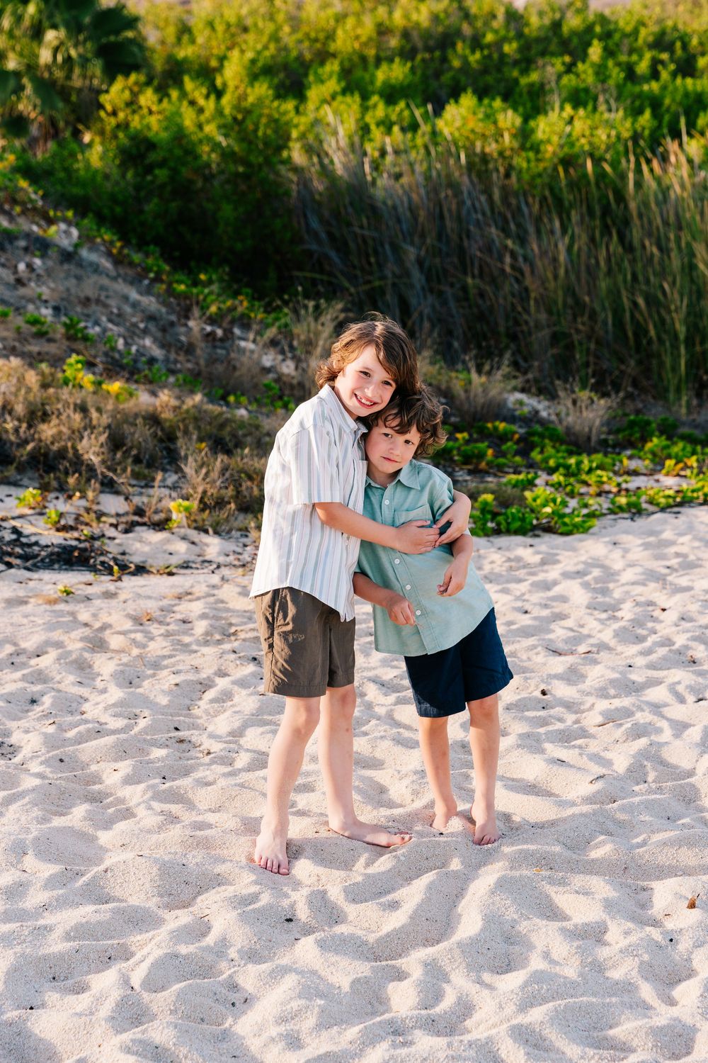 Two young boys hugging and smiling on a sandy beach with green coastal vegetation in the background.
