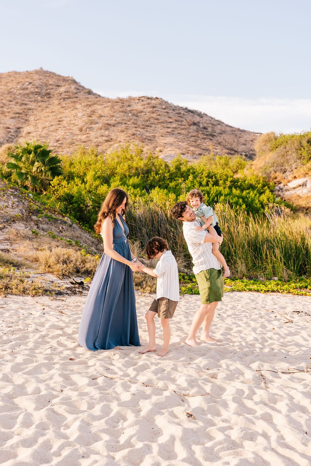 A pregnant woman in a blue dress enjoys a beach day with her family, including two children and a man, against a scenic hillside.