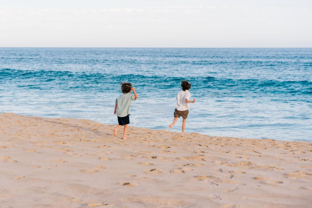 Two children running toward ocean waves on a sandy beach on a sunny day.