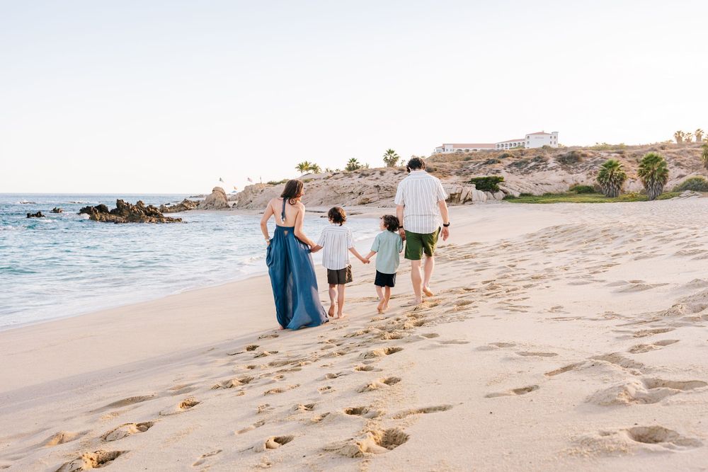 Family of four walking hand-in-hand along a sandy beach at sunset near rocky coastal cliffs.