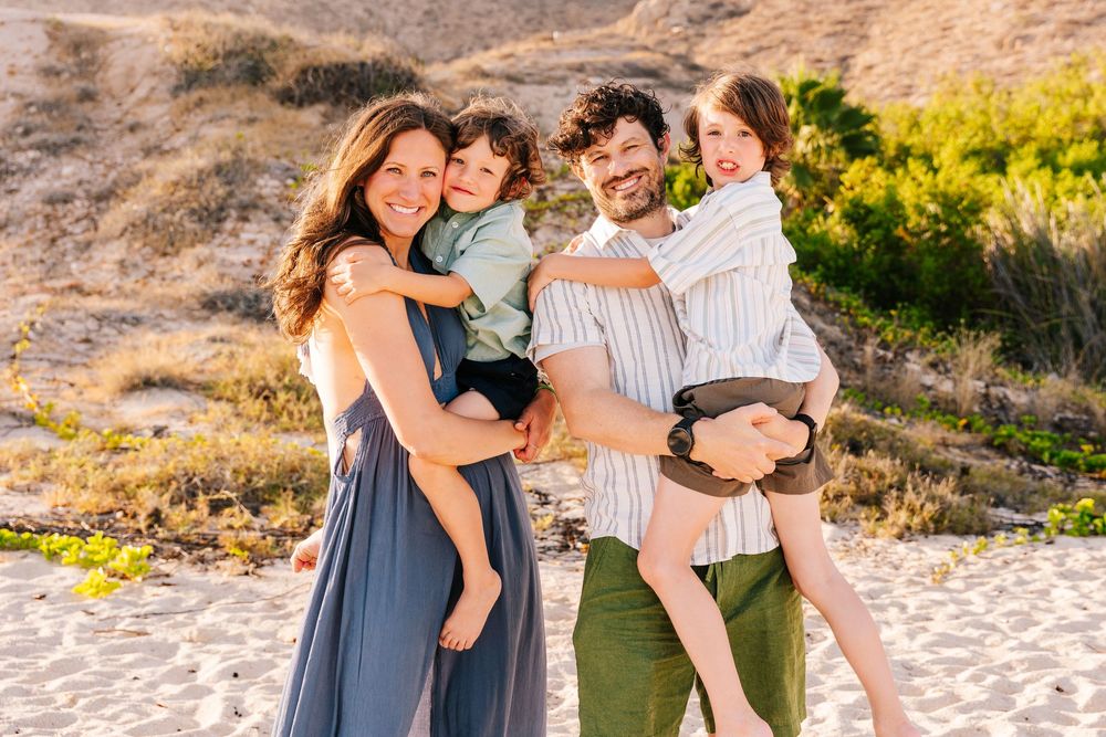 Happy family of four smiling on a sandy beach, parents holding young children with golden hills and greenery behind them.