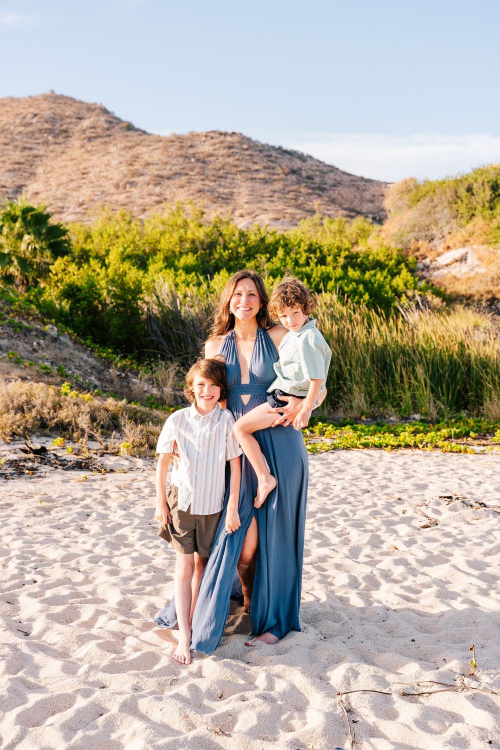 A mother in a blue maxi dress poses on a sandy beach with her two young sons, green hills in the background.