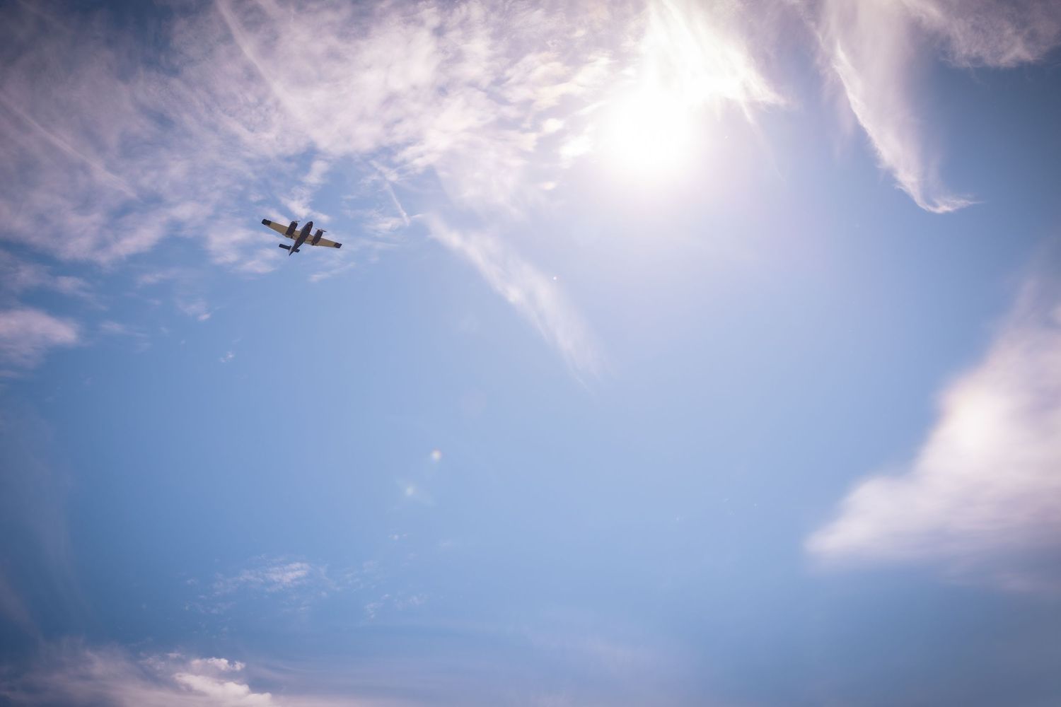 Small airplane flying through a bright blue sky with wispy white clouds and sunlight.