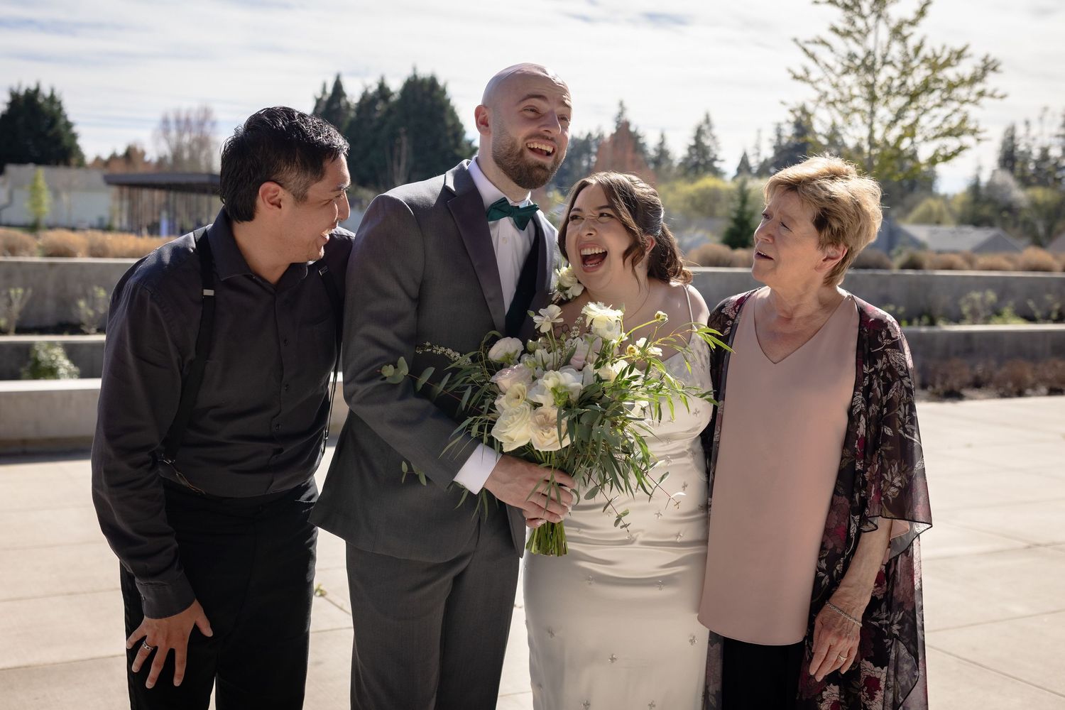 Bride and groom laughing with two family members outdoors, bride holding a white floral bouquet.