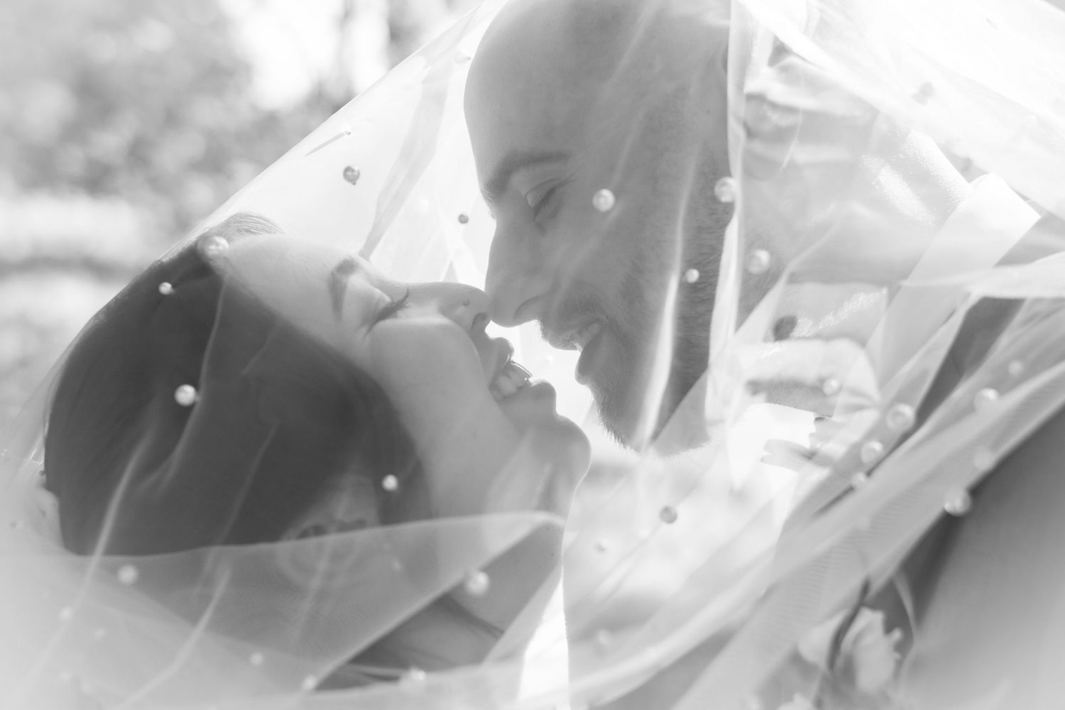 Black and white romantic photo of bride and groom nearly kissing beneath a pearl-dotted wedding veil.