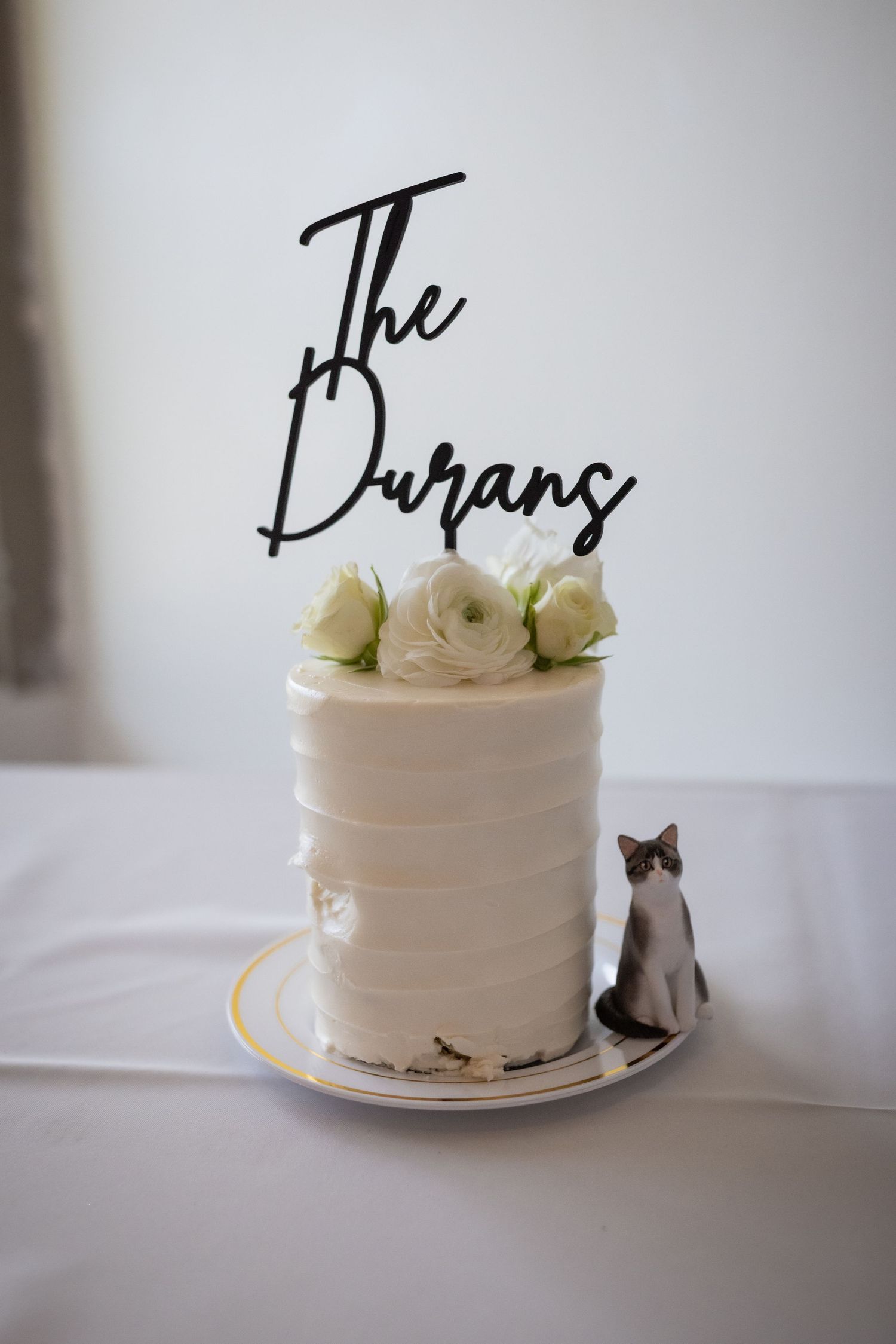 Elegant white wedding cake topped with white roses and a black acrylic topper reading The Durans.