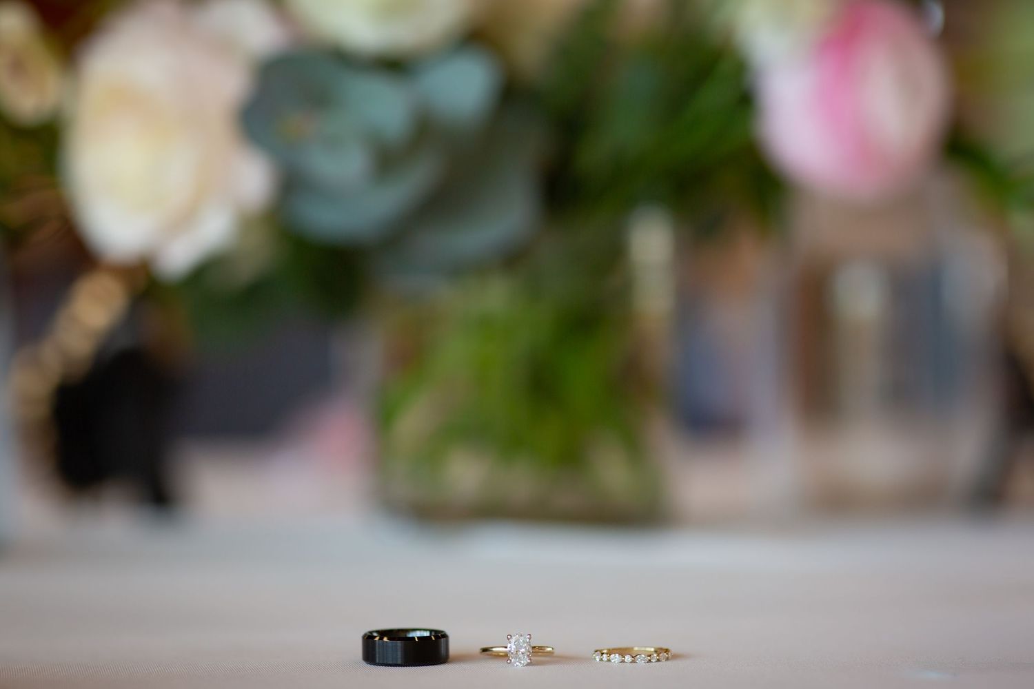Black wedding band and gold earrings displayed on white surface with soft floral arrangement in background.