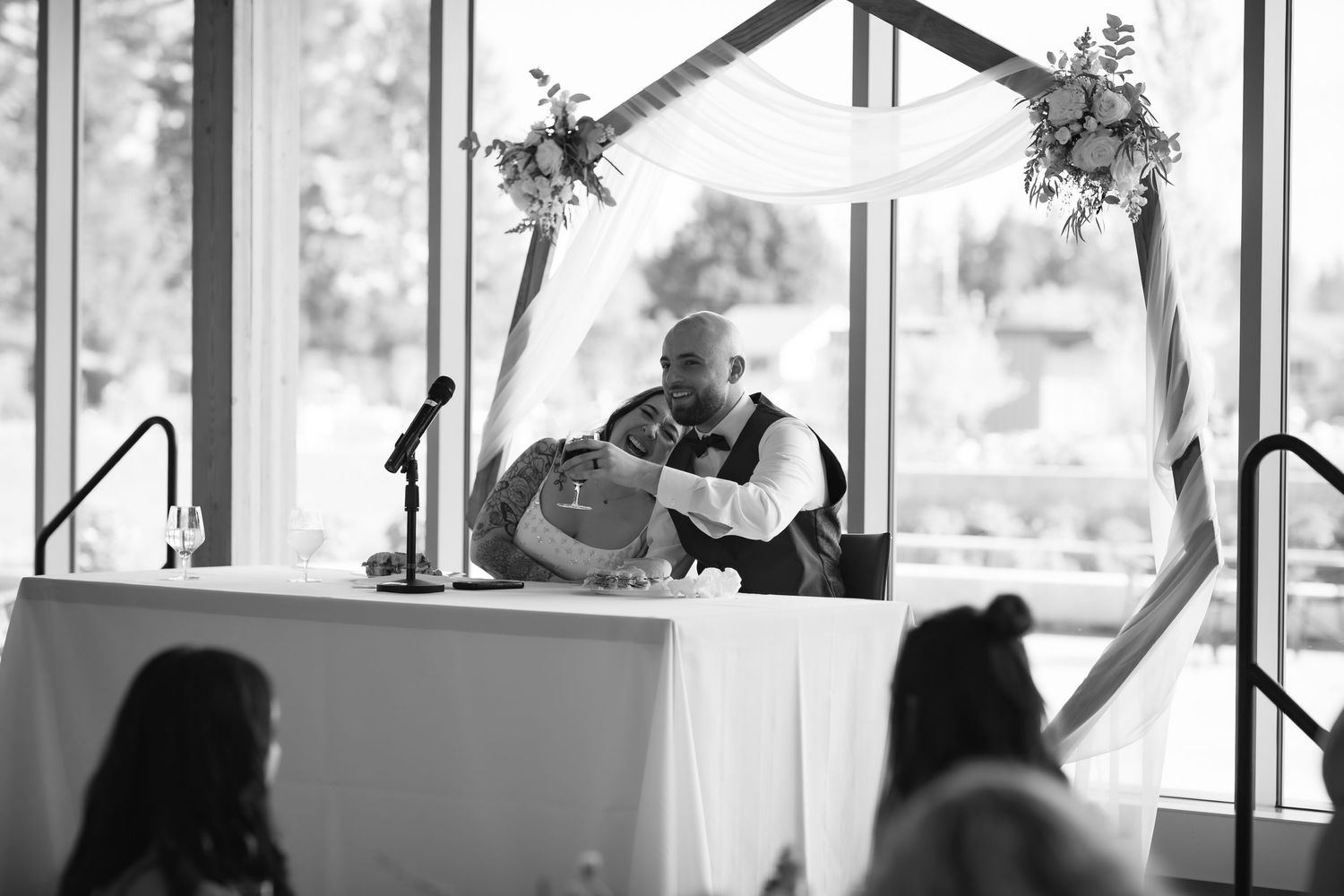 Groom seated at altar table beneath floral arch during indoor wedding ceremony in black and white.
