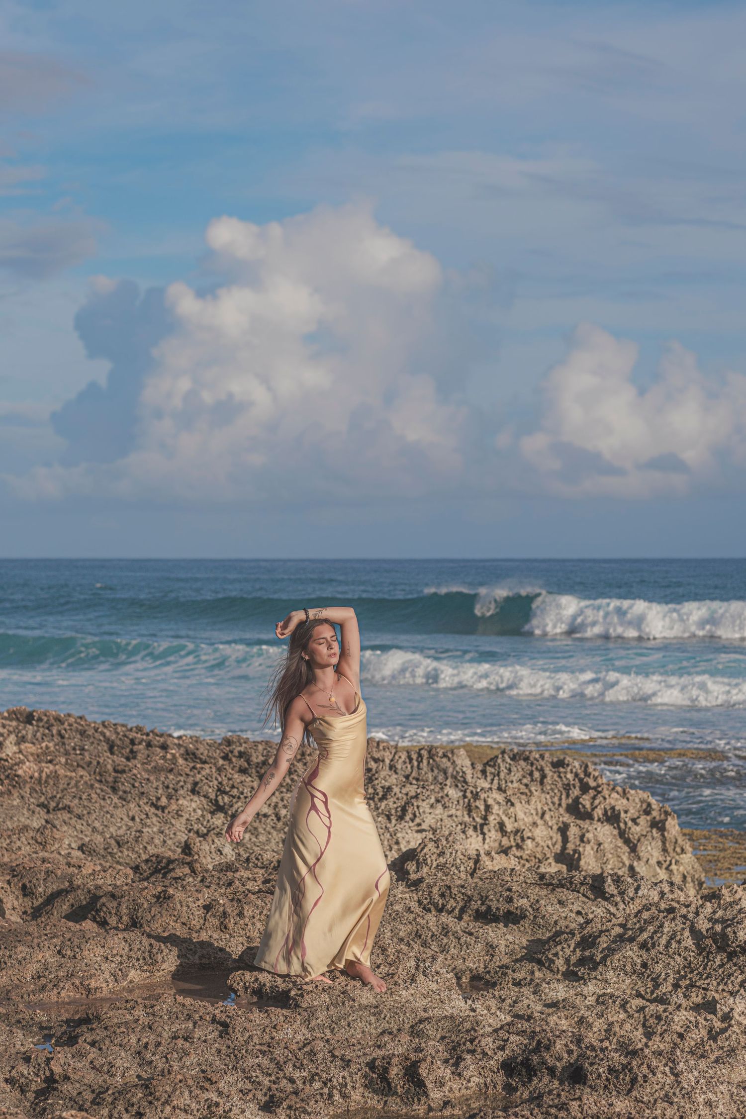 Woman in a flowing dress photographed along Oahu's North Shore coastline with ocean waves and dramatic skies during a lifestyle shoot.