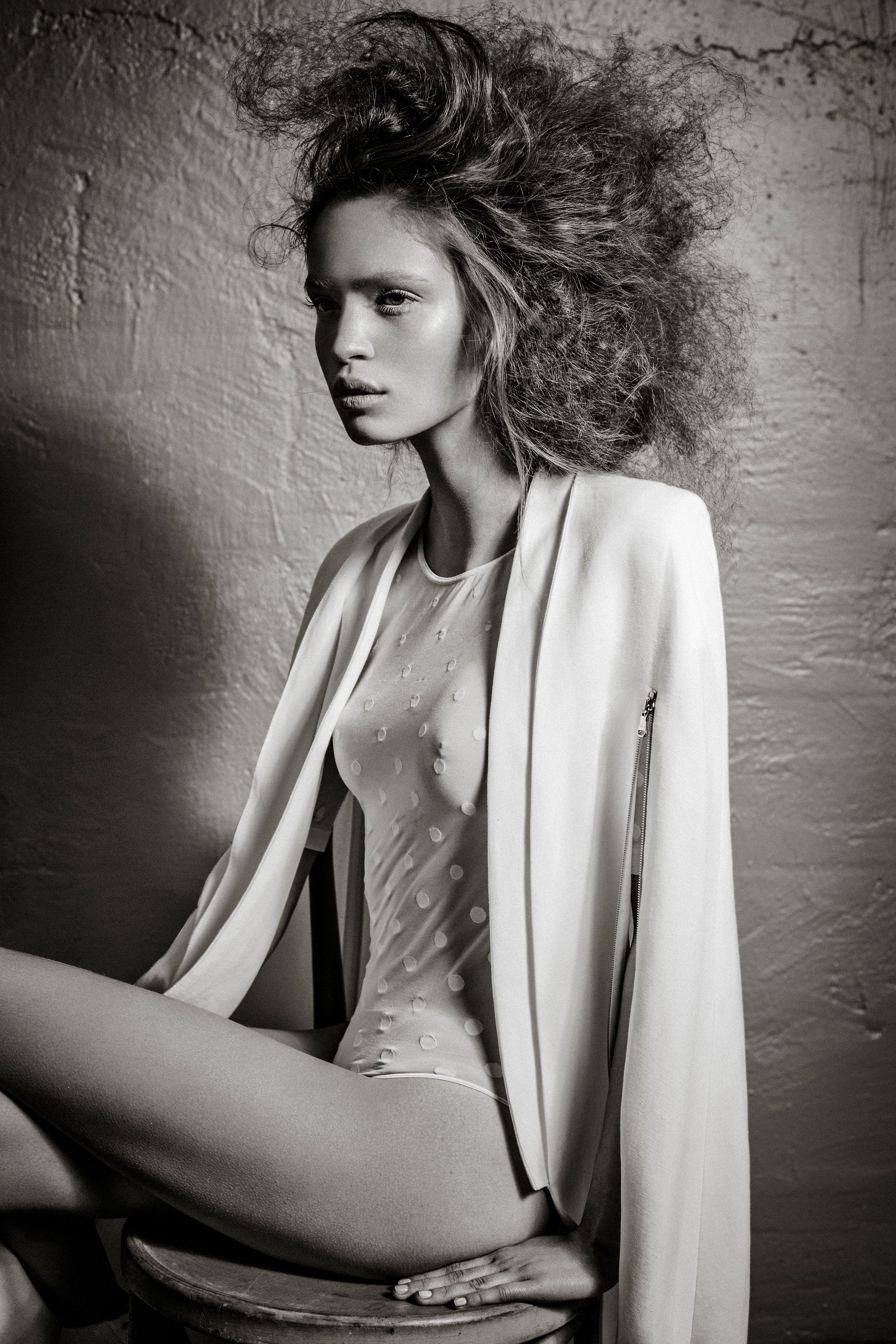 Black and white fashion photo of a woman with voluminous hair wearing a white blazer and sheer bodysuit.