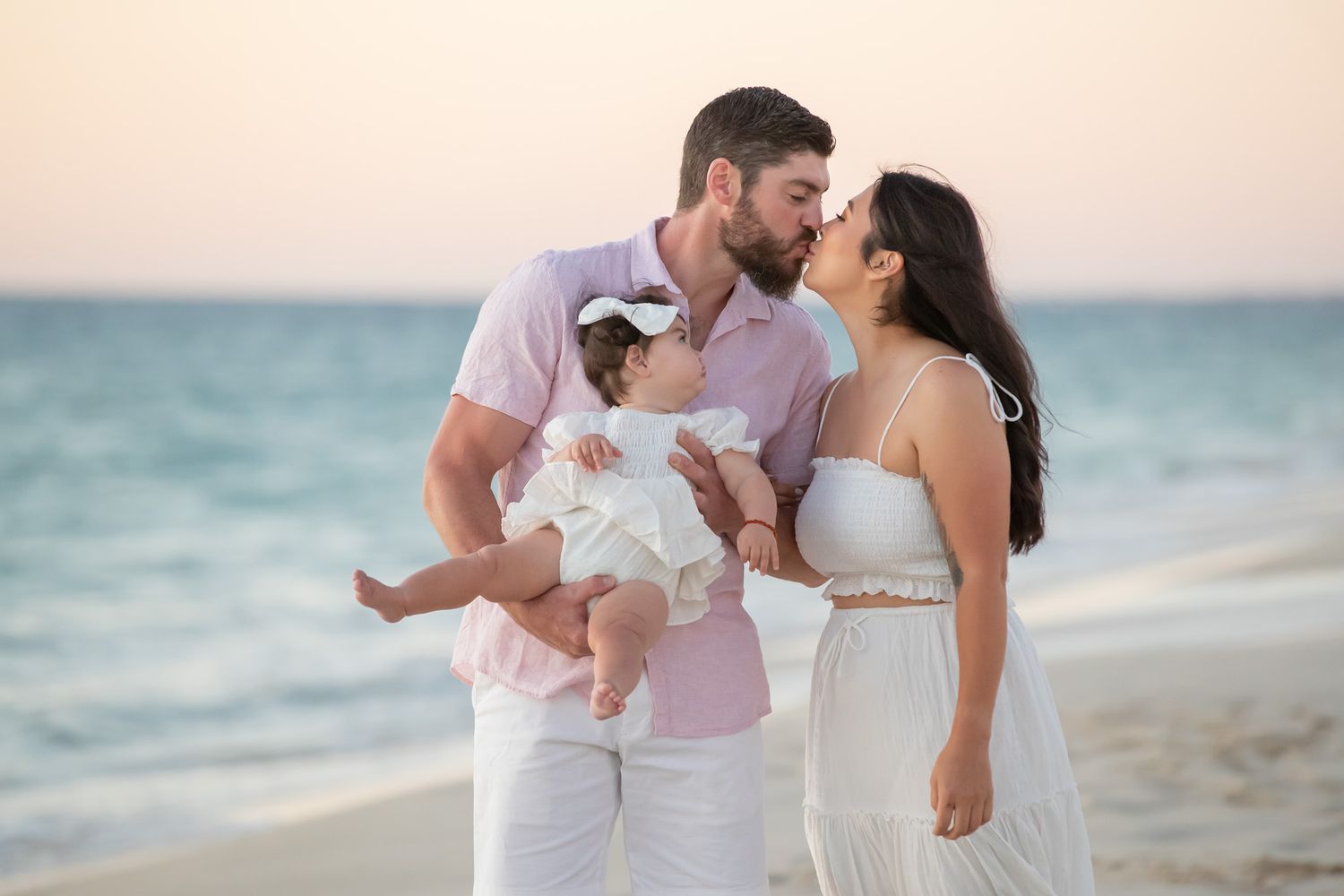 Parents kissing over baby girl on sandy beach at dusk, family dressed in white and pink.