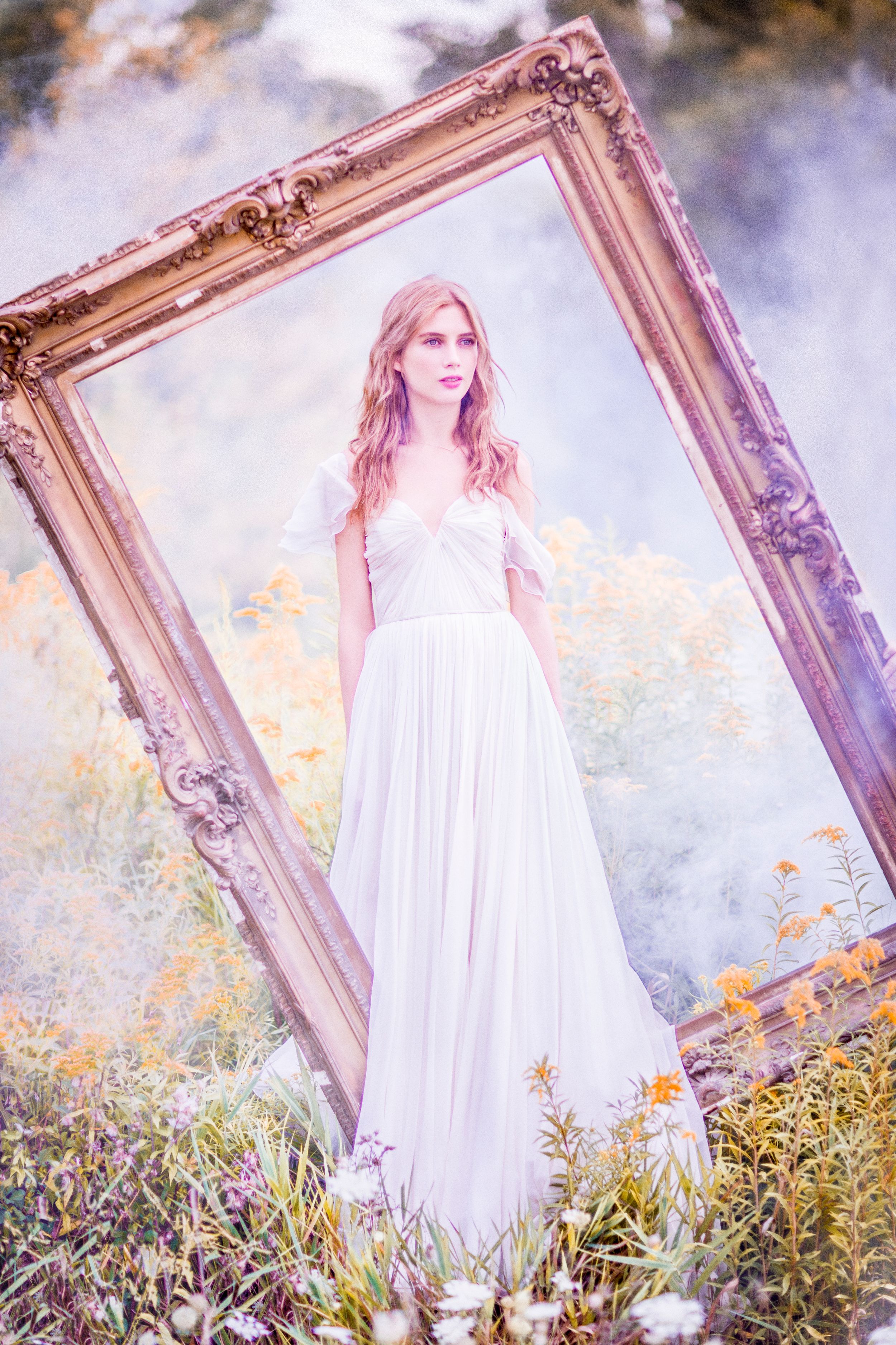 Woman in white gown stands within ornate gold frame surrounded by wildflowers in dreamy, soft-focus fantasy setting.