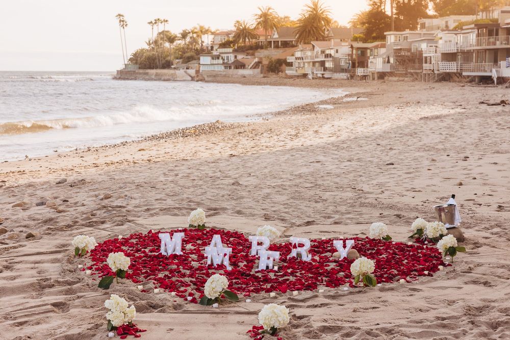 Rose petal marriage proposal spelling MARRY on a sandy beach at sunset with white flowers and coastal homes in background.
