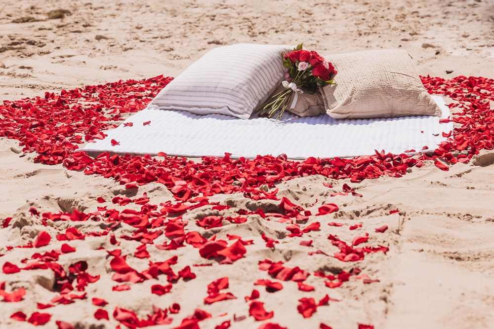 Red rose petals arranged in a heart shape on sandy beach with white blanket, pillows, and bouquet for romantic occasion.