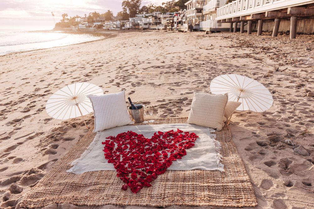 Romantic beach setup with rose petal heart, white umbrellas, cushions, and champagne on a rug at sunset.