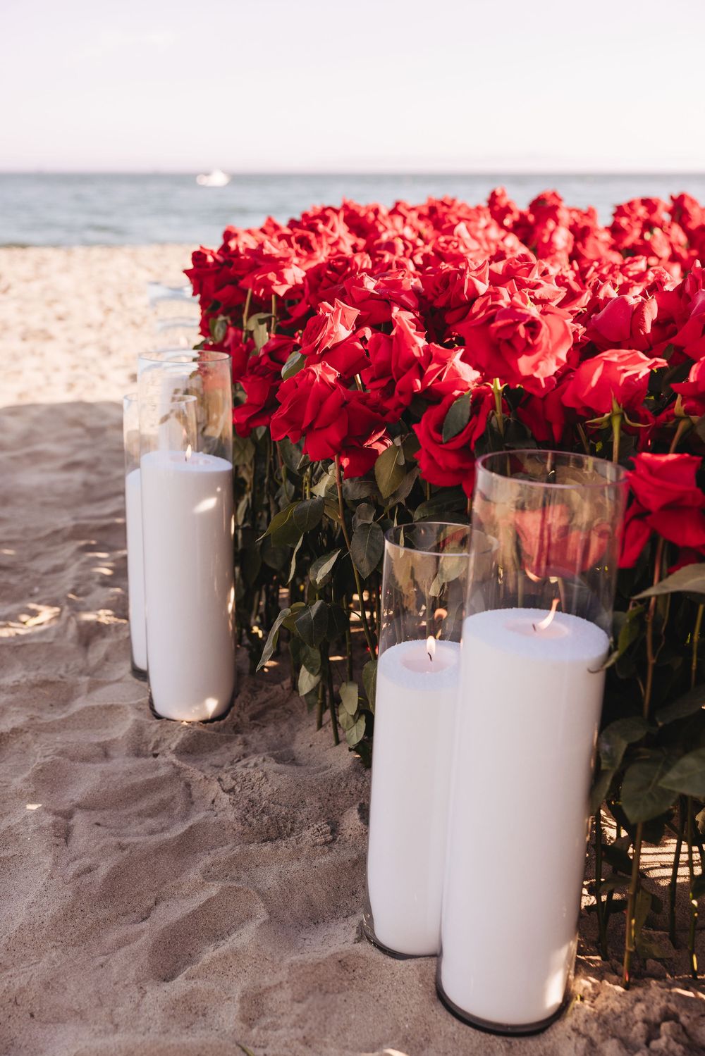 Red roses and white pillar candles in glass holders arranged on a sandy beach with ocean in the background.