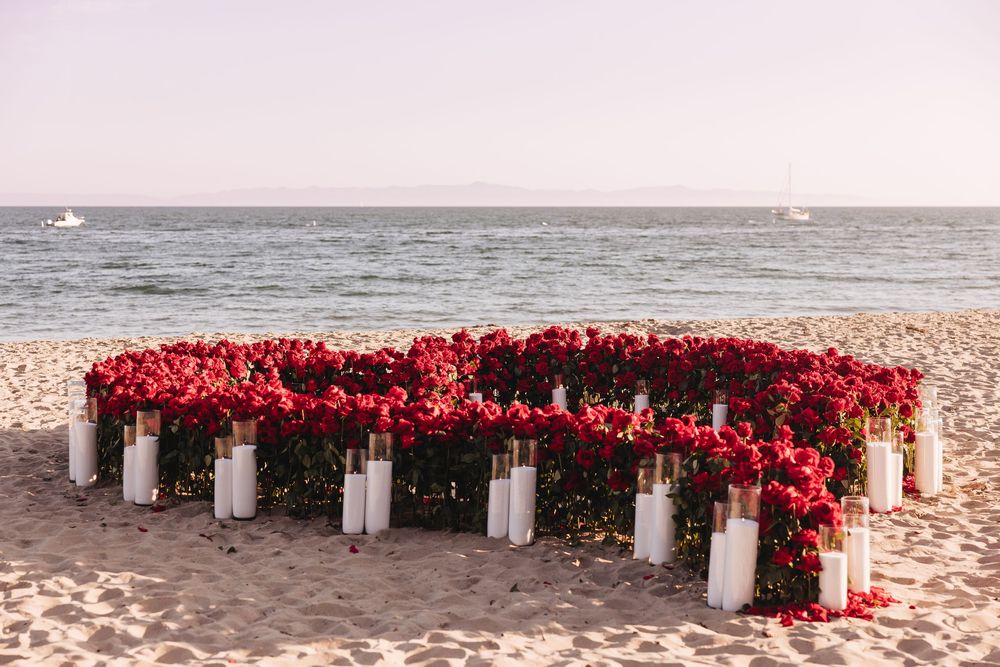 Heart-shaped red rose arrangement with white pillar candles on a sandy beach with ocean views.
