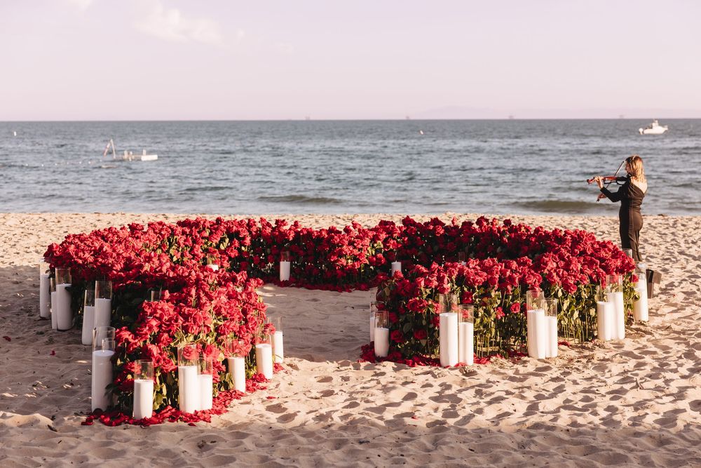 Heart-shaped red rose arrangement with white candles on a sandy beach, romantic proposal setup with ocean view.