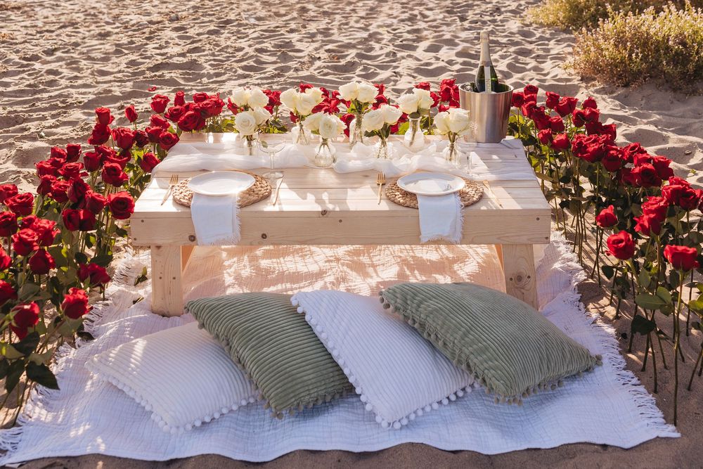 Romantic beach picnic setup with red and white roses, wooden table, white blanket, and green pillows on sandy shore.
