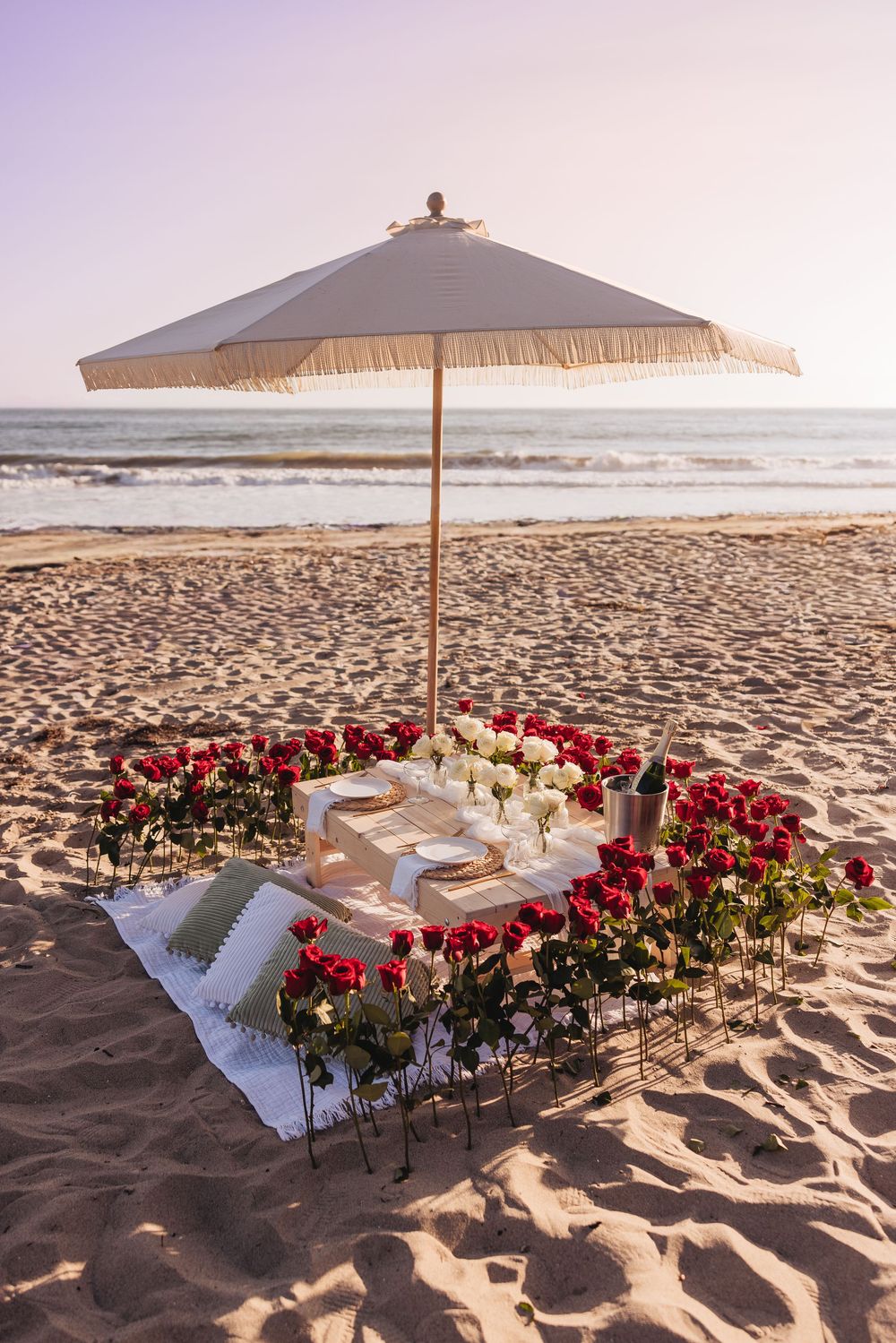 Romantic beach picnic setup with red and white roses, elegant table setting, and fringed umbrella on sandy shore at sunset.