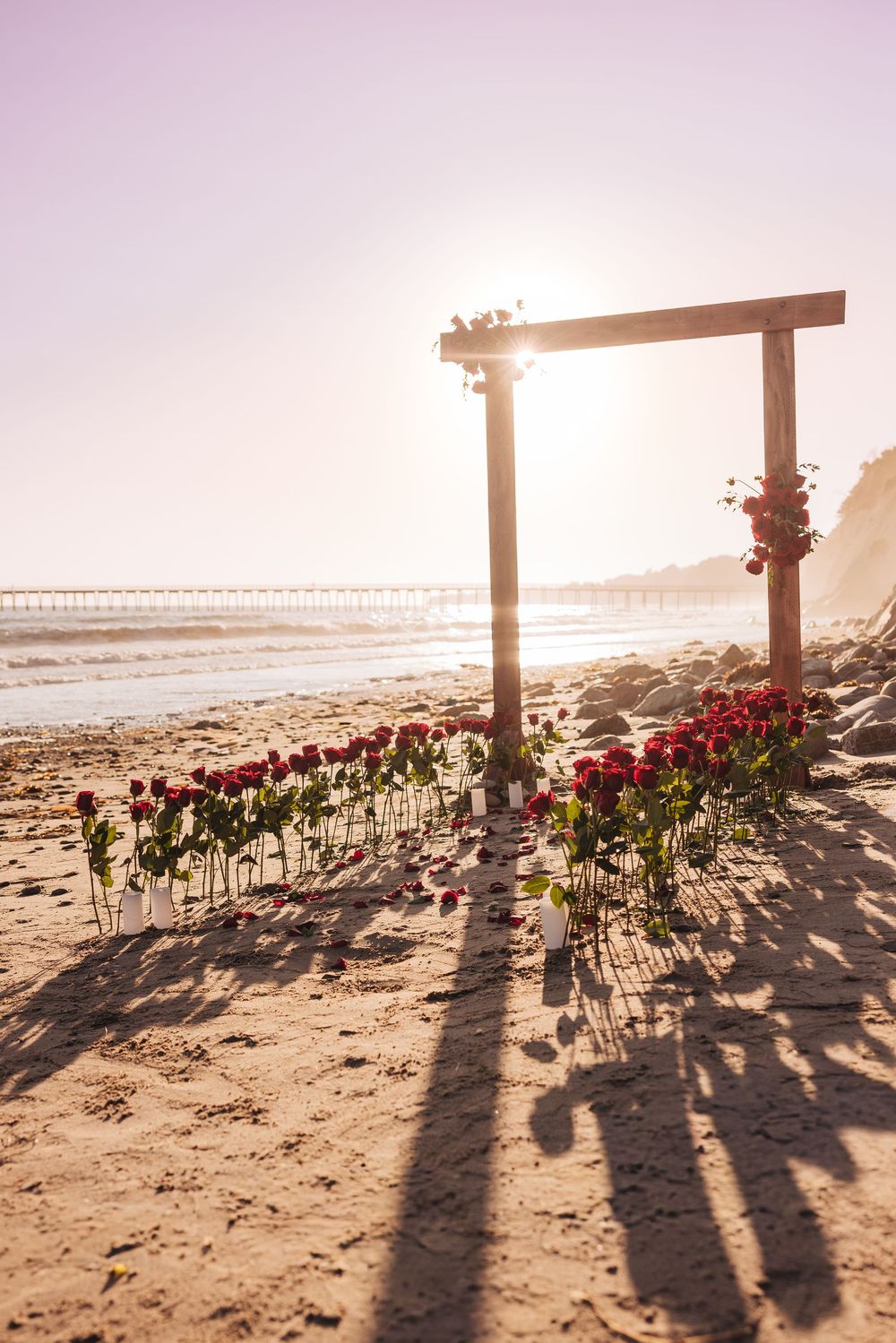 Romantic beach wedding arch with red roses lining the aisle at sunset, ocean pier visible in the background.