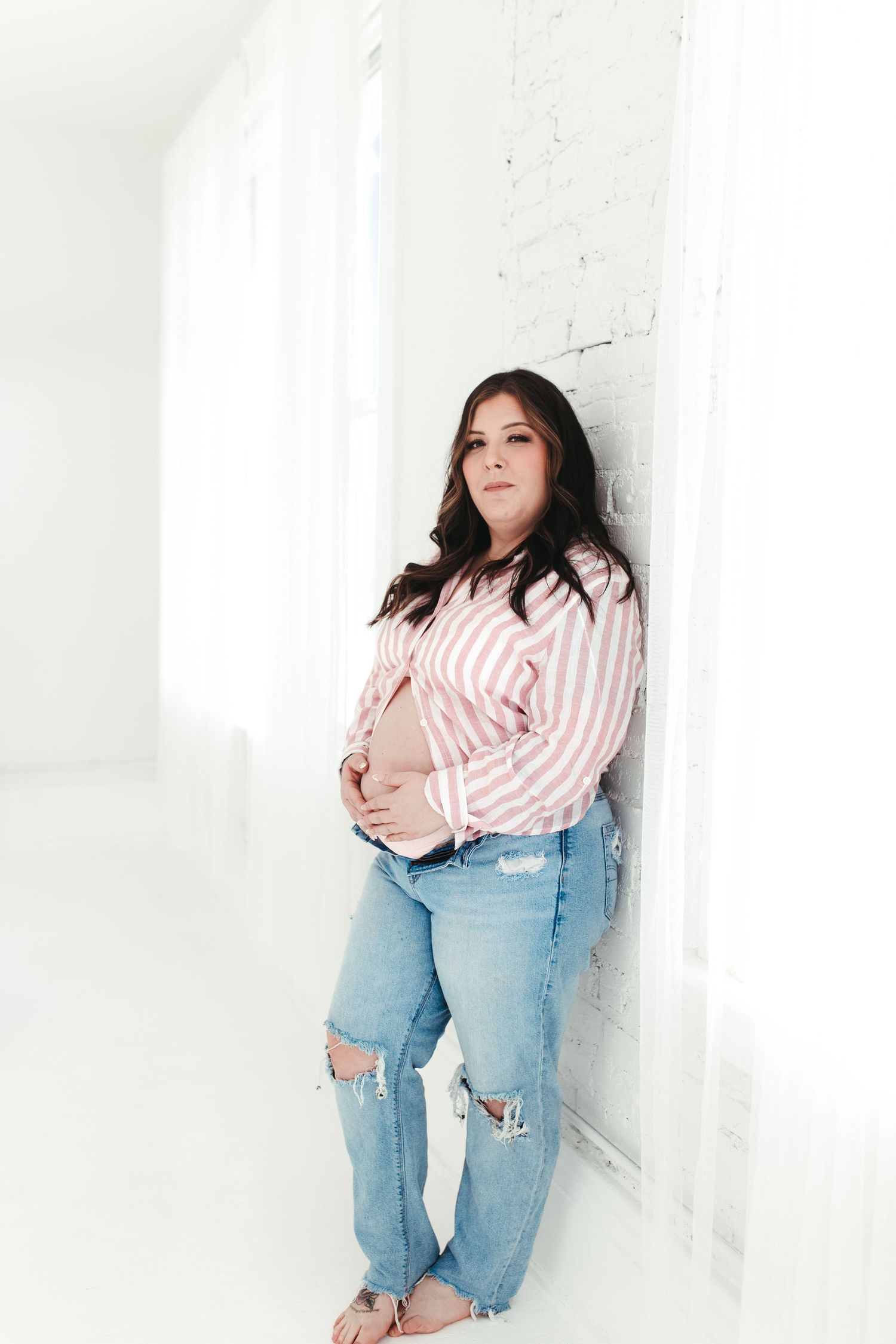 Pregnant woman in pink striped shirt and ripped jeans holding baby bump against white brick wall.