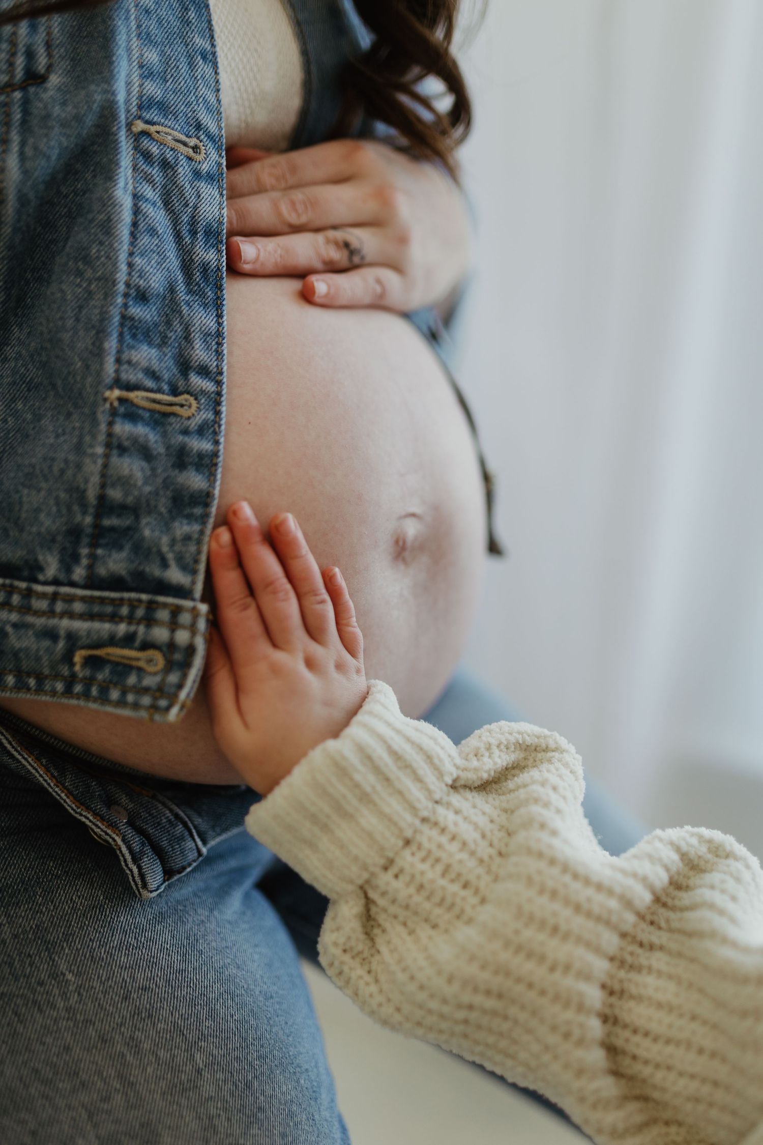 Close-up of child's hand gently touching pregnant mother's bare belly in denim jacket.