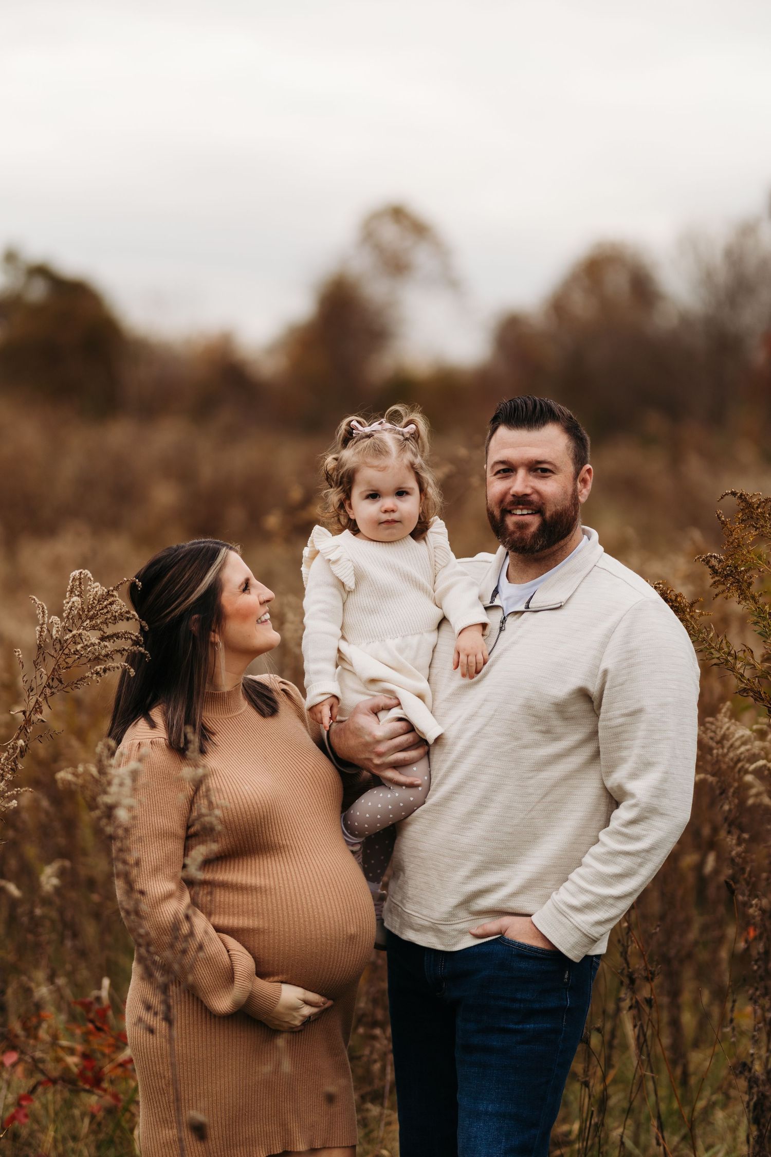 A smiling pregnant couple poses with their toddler daughter in a golden autumn field for a maternity photo.