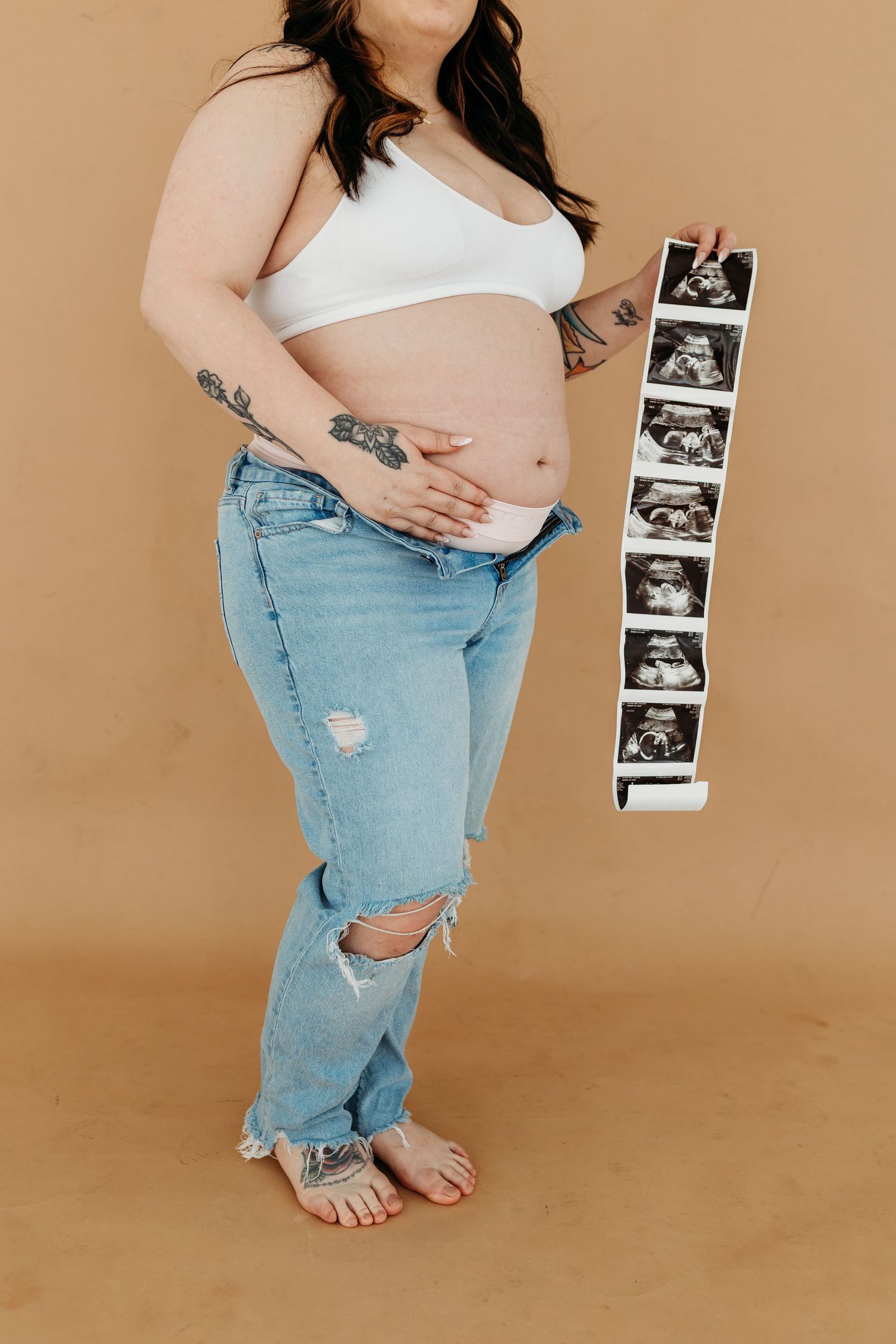 Pregnant woman with tattoos in white bralette and jeans holding ultrasound strip on beige background.