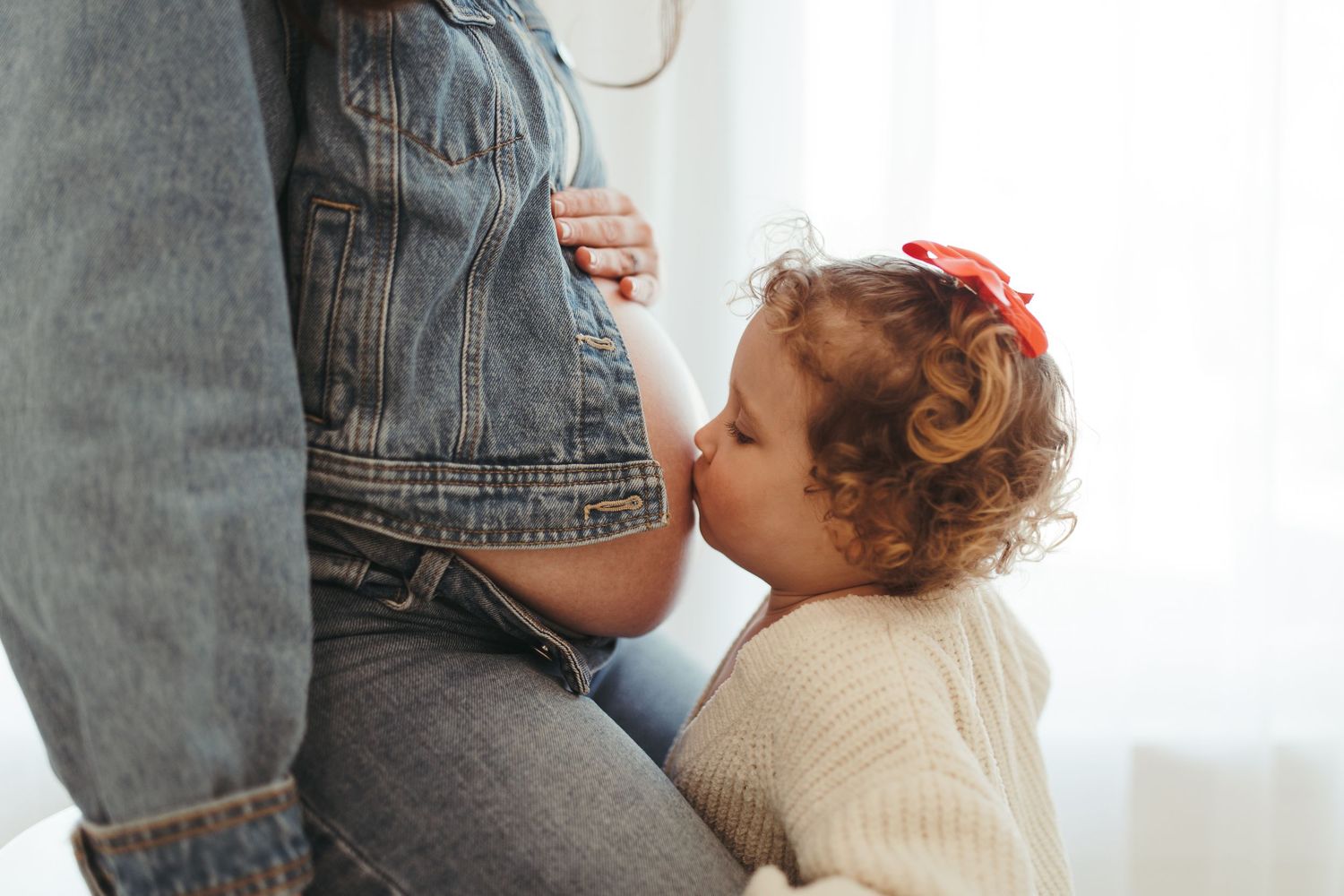 Curly-haired toddler in cream sweater kissing pregnant mother's bare belly tenderly.