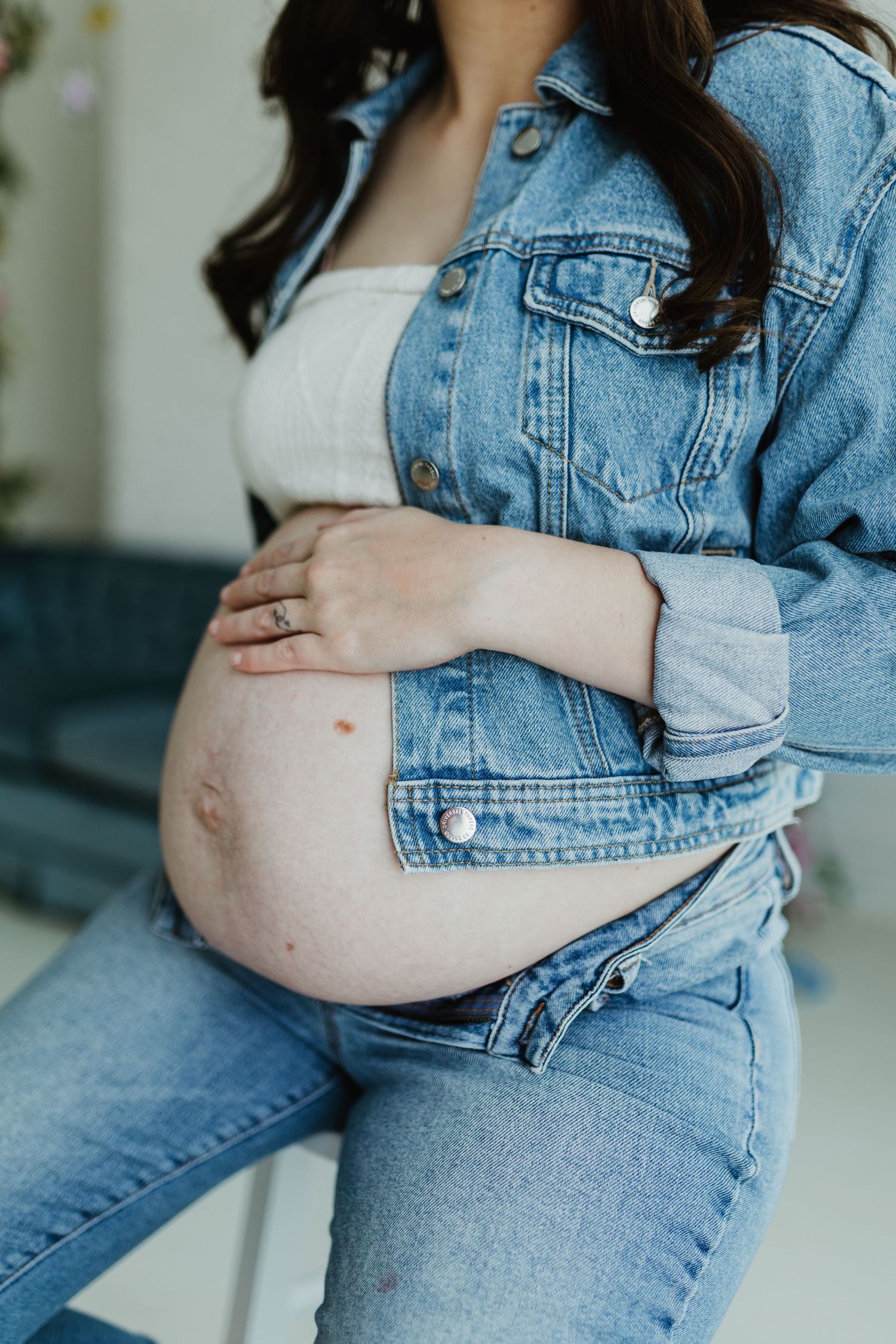 Close-up of pregnant woman in denim jacket cradling her bare baby bump while seated.