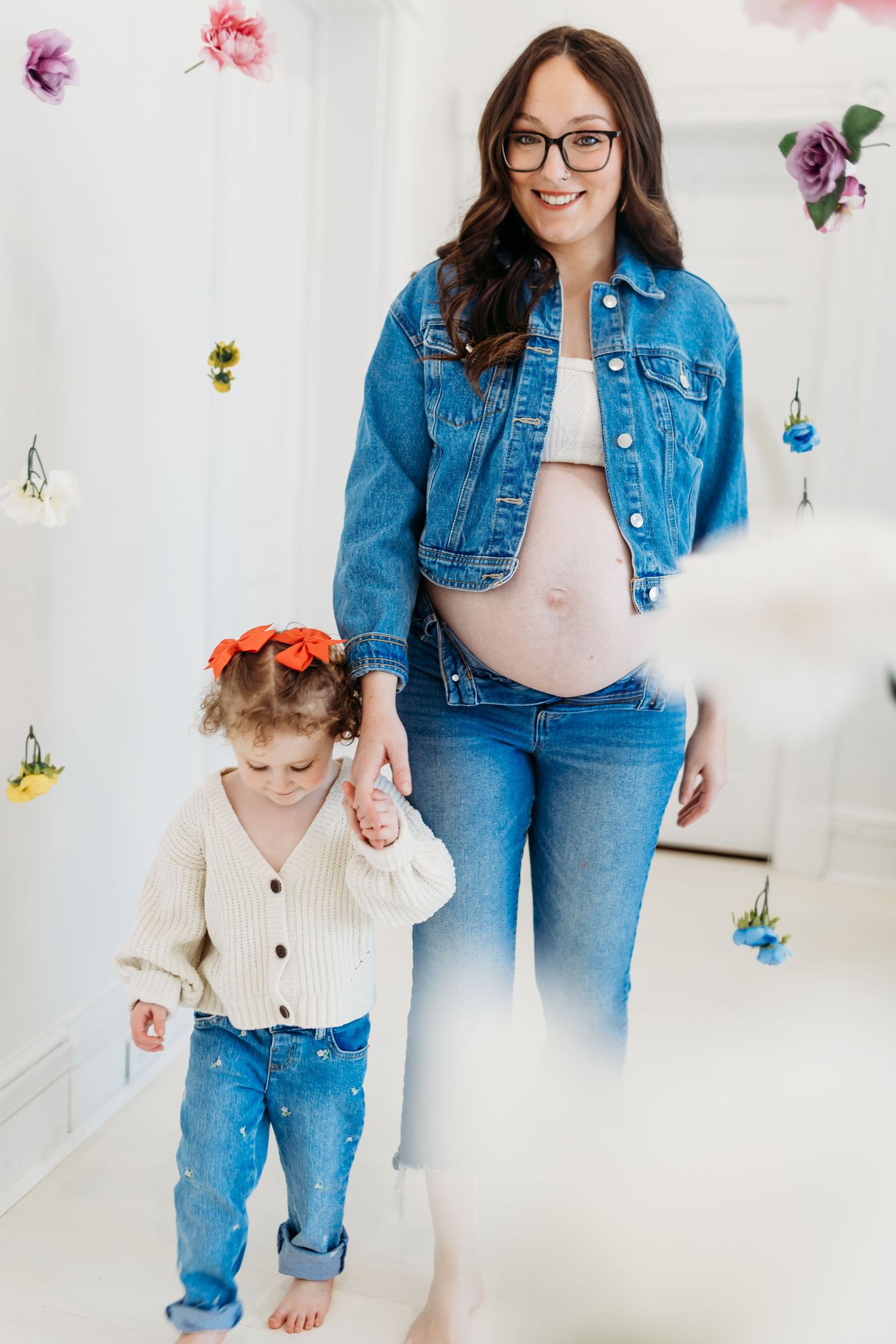 Smiling pregnant woman in glasses and denim jacket holding toddler's hand amid hanging flowers.