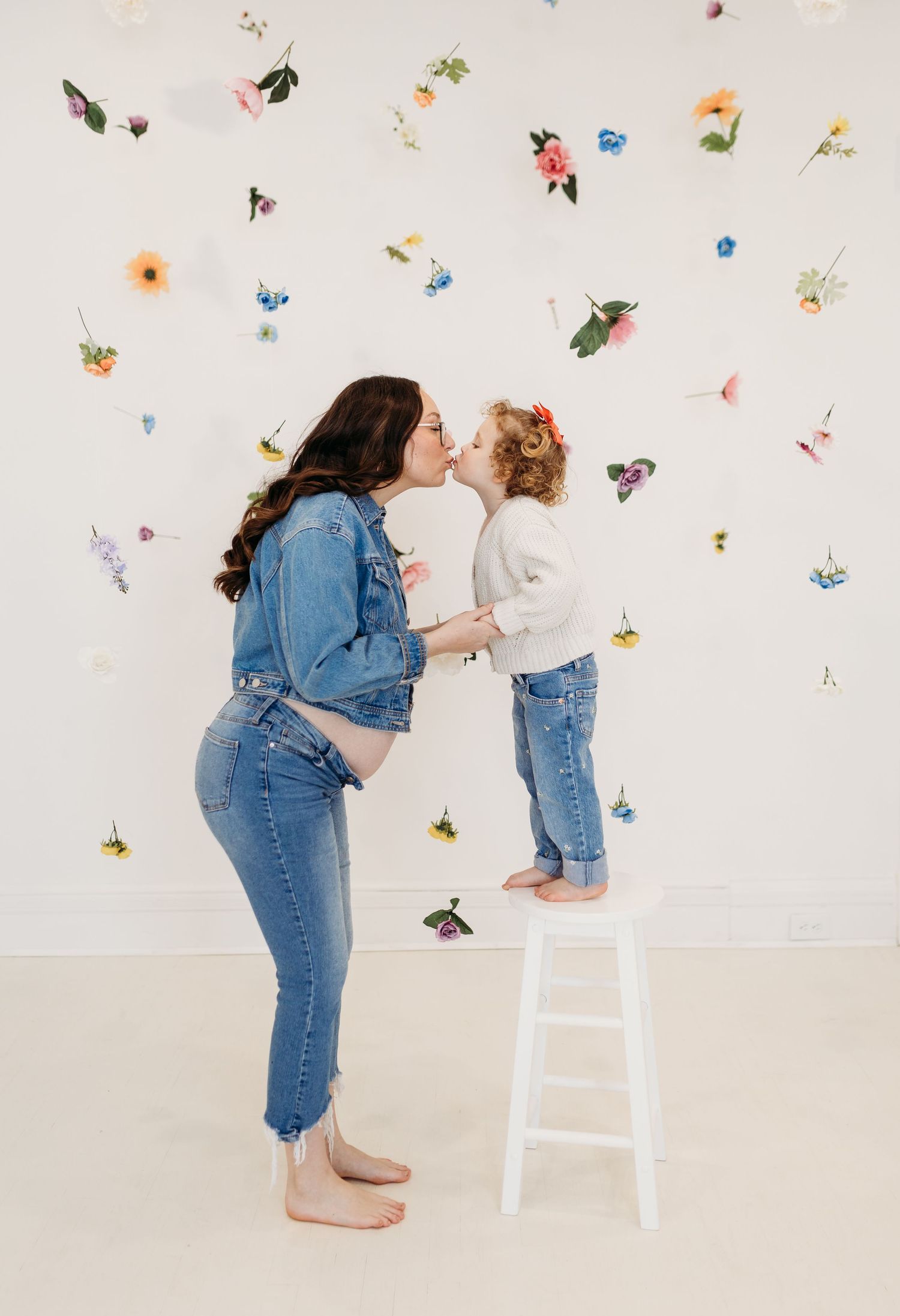 Pregnant mom and toddler sharing a kiss, standing before a floral wall backdrop.