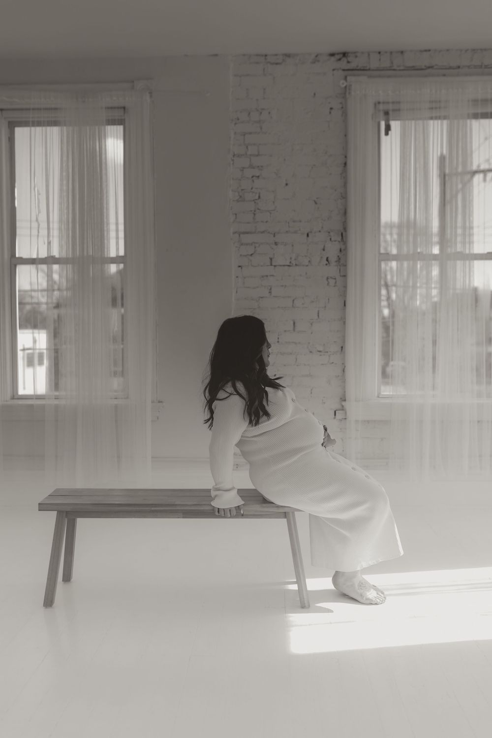 Black and white photo of pregnant woman in white dress sitting on wooden bench near windows.