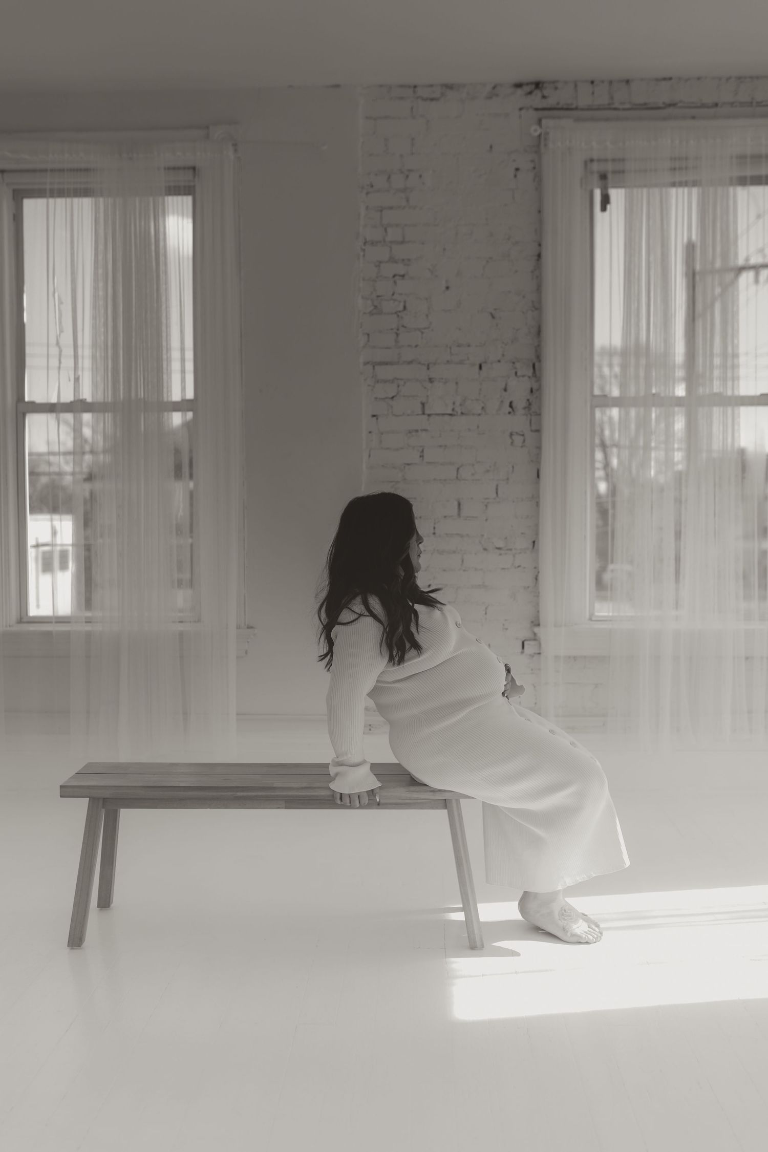 Black and white photo of pregnant woman in white dress sitting on wooden bench near windows.