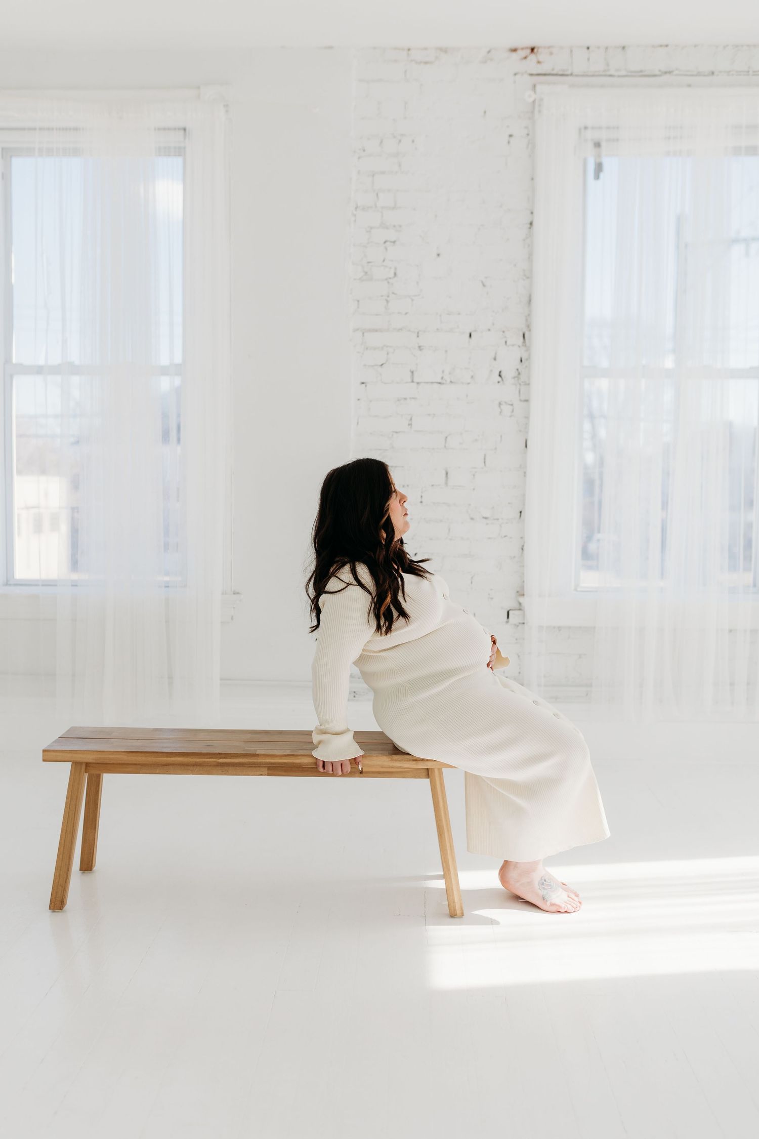 Pregnant woman in cream knit dress sitting on wooden bench in bright white studio with brick walls.