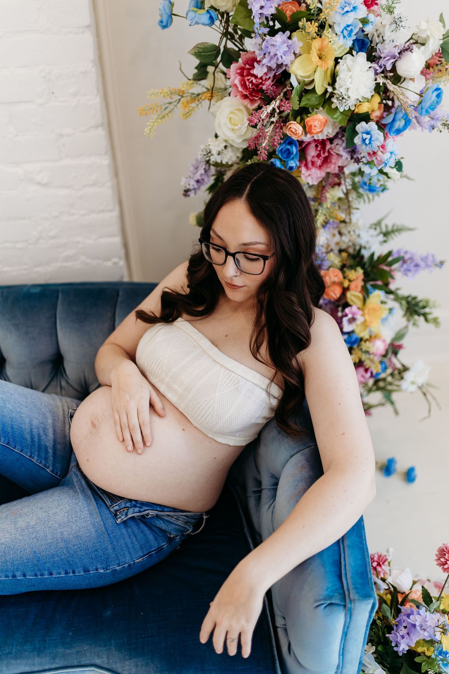 Pregnant woman in glasses resting on blue velvet chair beside colorful floral arrangement.