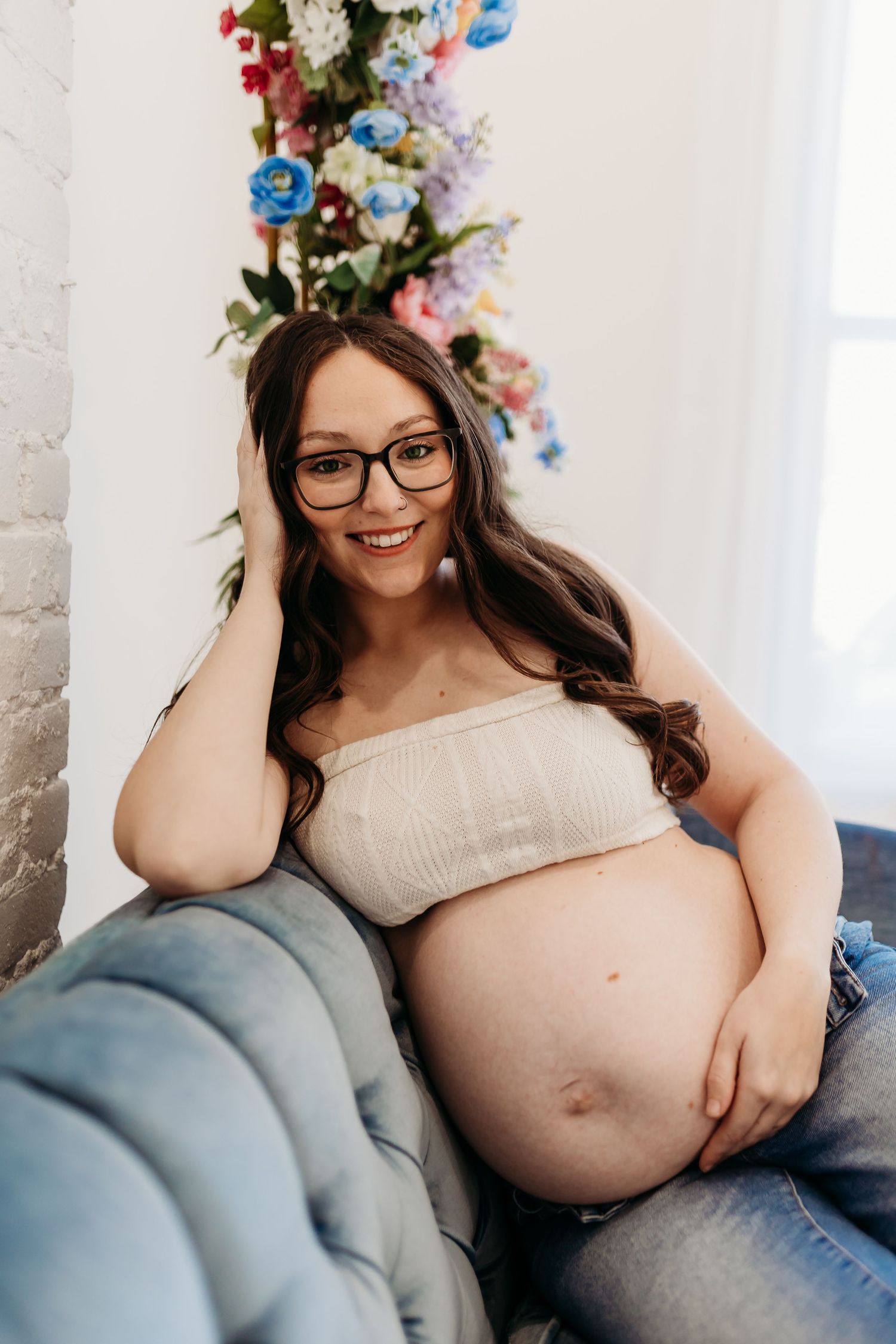 Smiling pregnant woman in glasses and white bandeau top relaxing on blue velvet sofa.