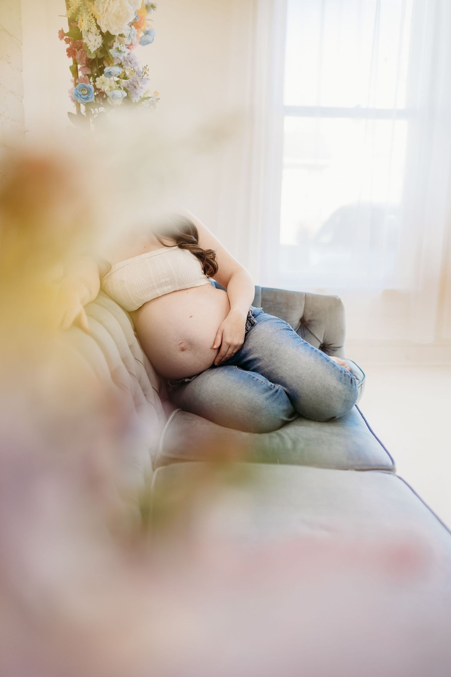 Pregnant woman curled up resting on gray sofa near bright window with soft light.