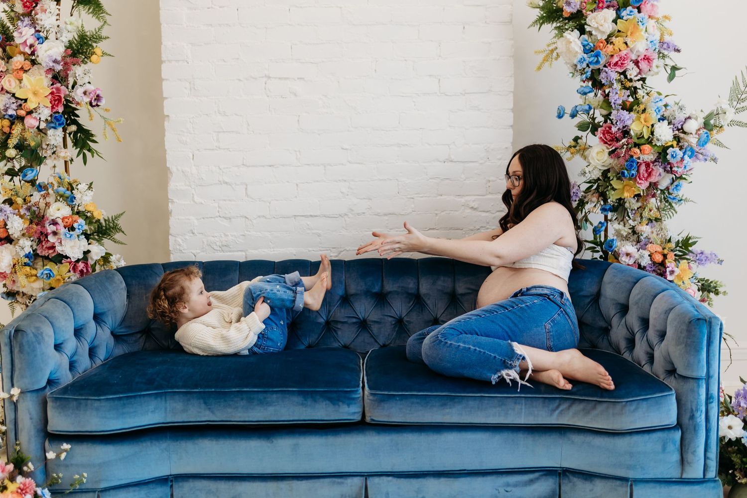 Mother and toddler playing on a blue velvet sofa surrounded by colorful floral arrangements in a bright studio.