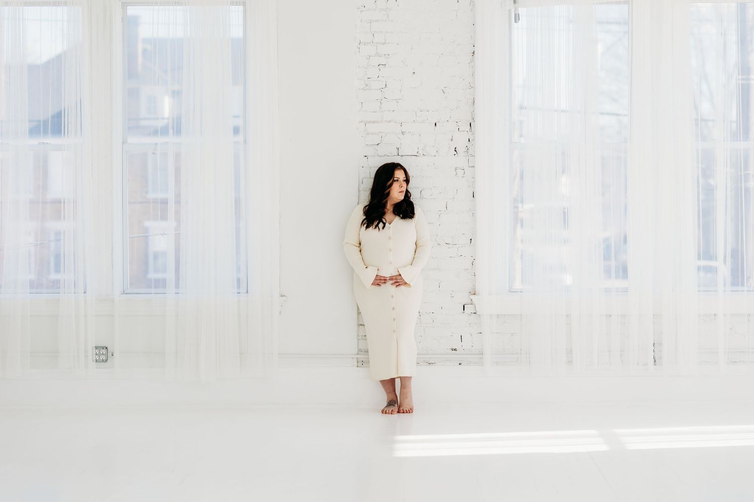 Pregnant woman in white lace dress standing in bright white studio with large windows.