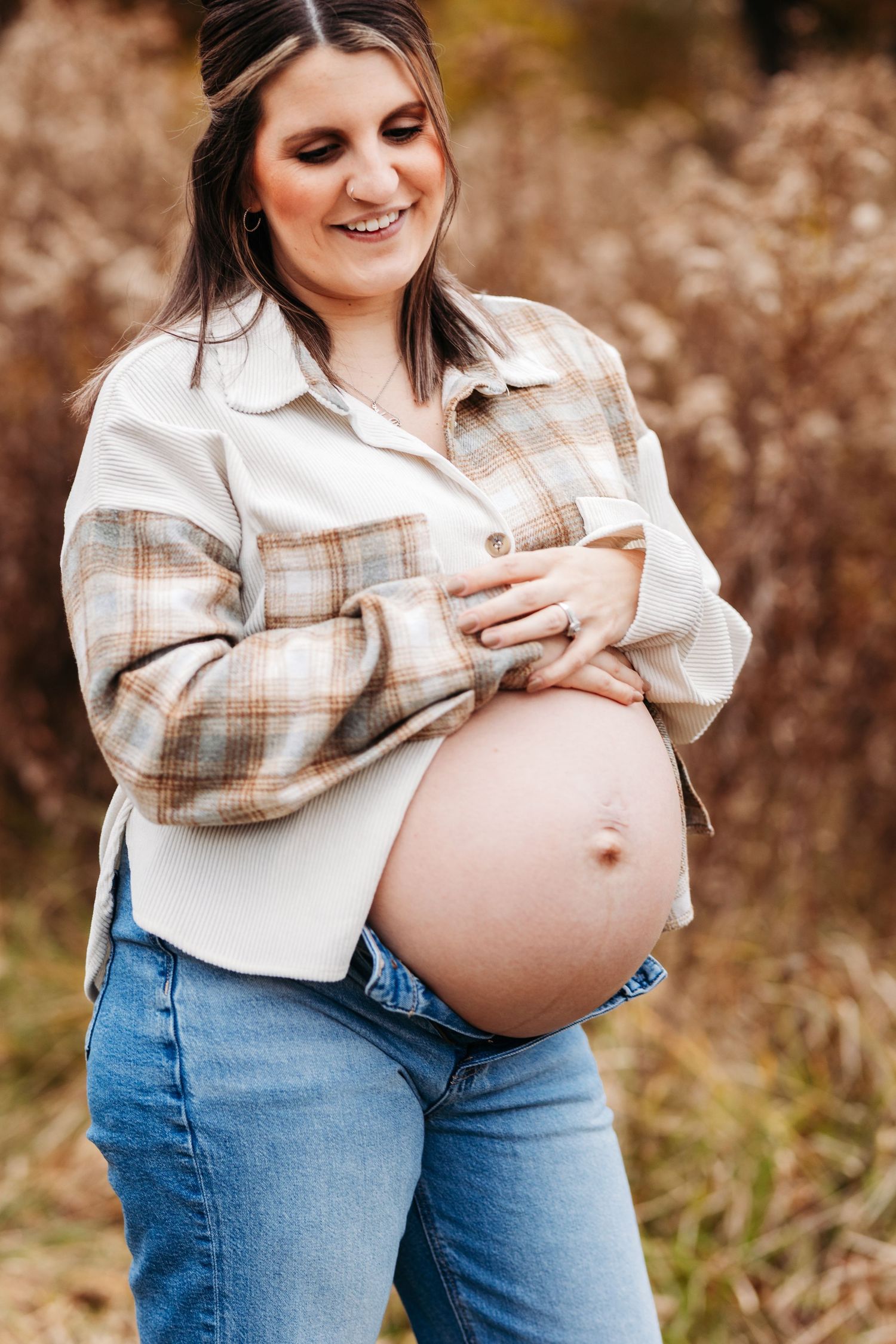 A smiling pregnant woman in a plaid shirt and jeans cradles her bare baby bump in an autumn field.