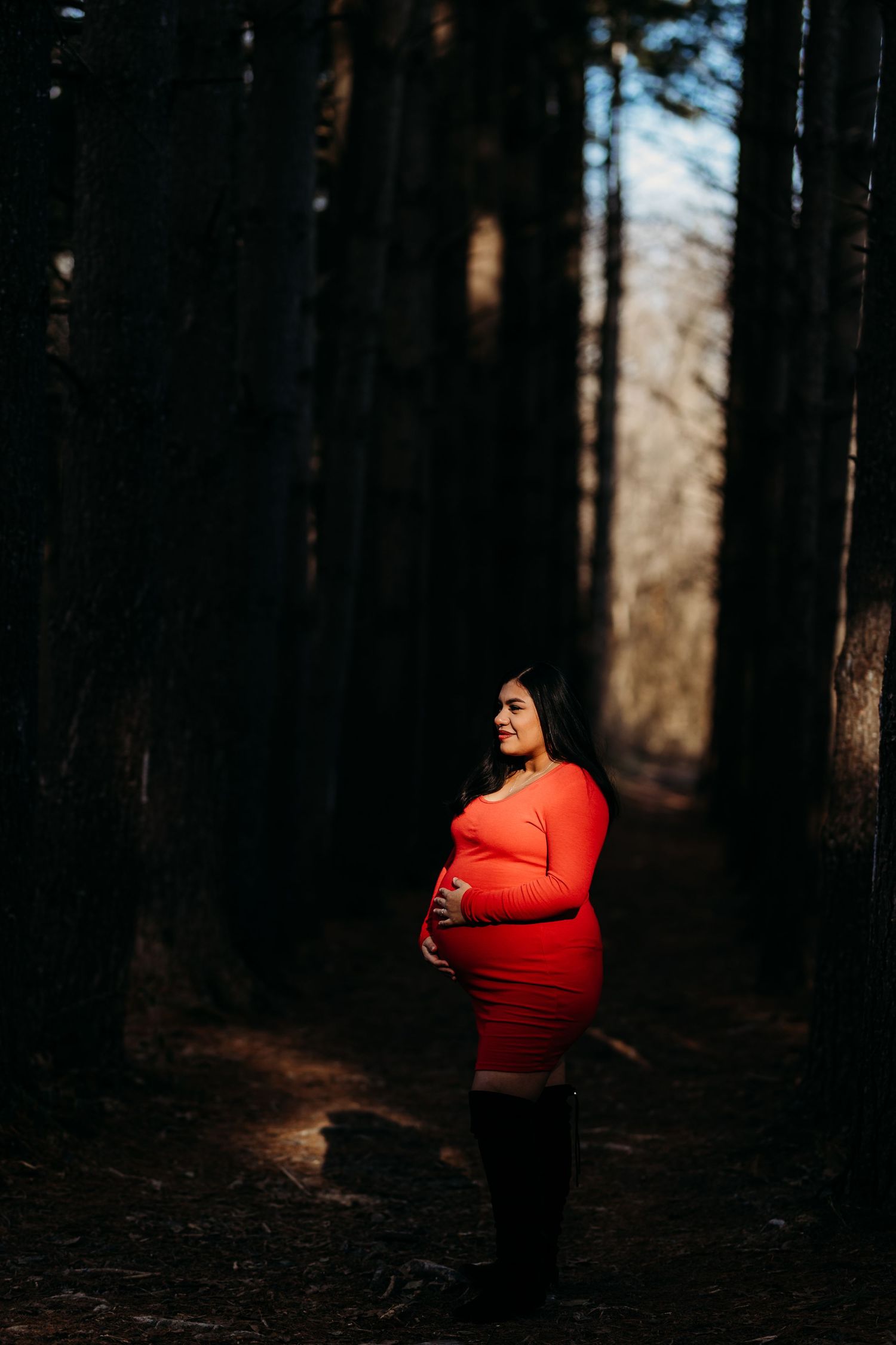 Pregnant woman in red dress cradling baby bump on a dramatic shadowy forest path.