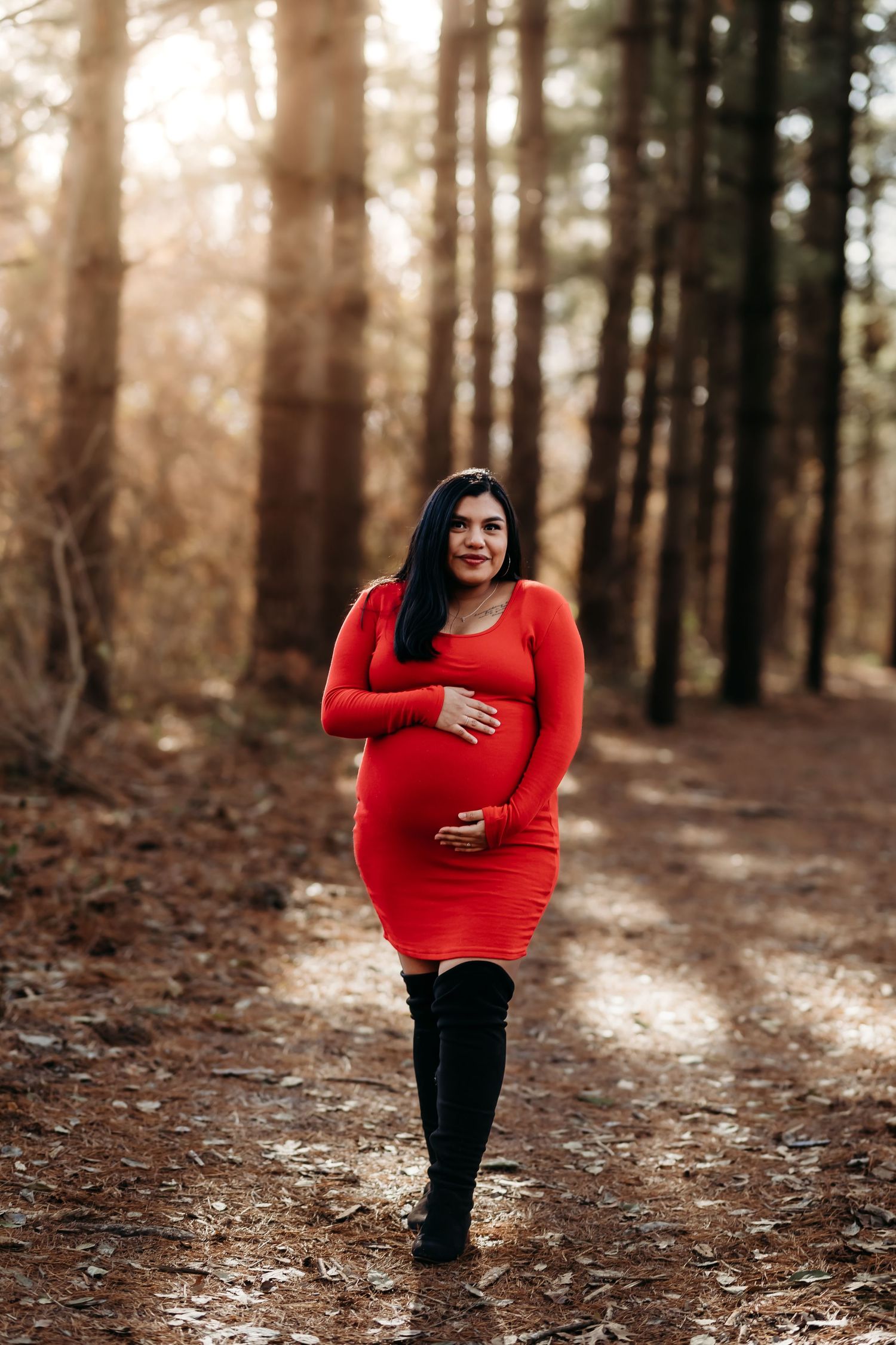 Pregnant woman in red dress and black boots walking on a sunlit forest path.