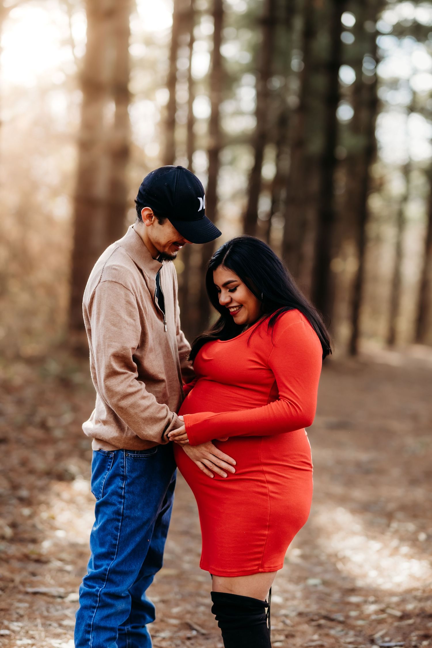 Couple smiling and looking down at pregnant belly on a sunlit forest path maternity shoot.