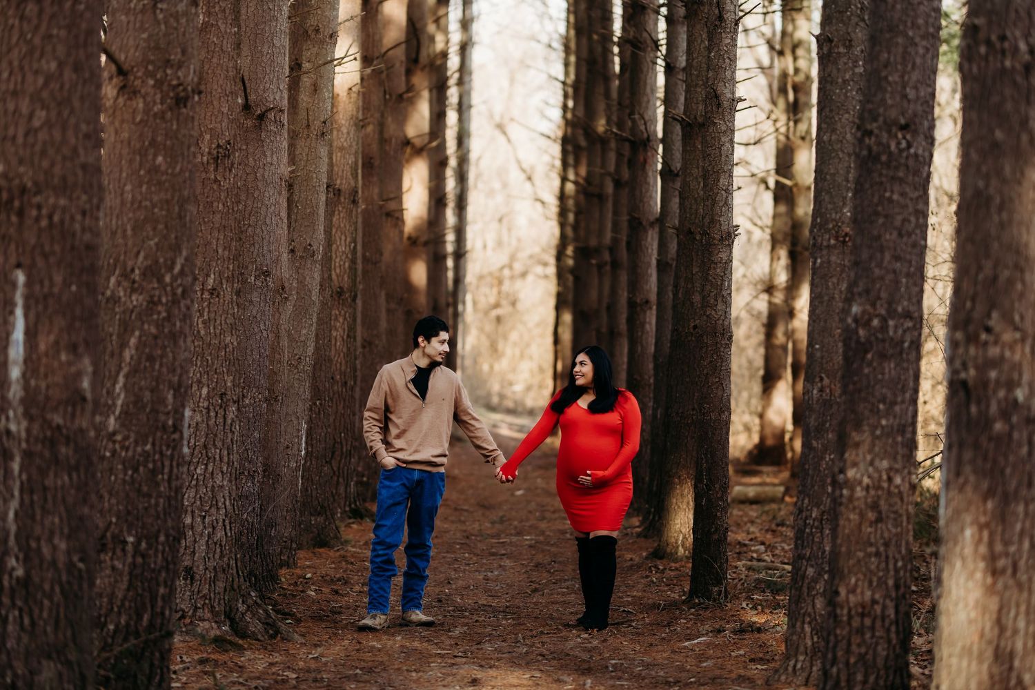 Couple holding hands walking through tall pine trees in a forest, woman wearing red dress during maternity photo session.