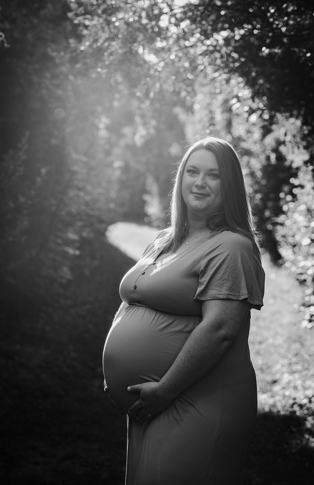Black and white portrait of smiling pregnant woman in maxi dress holding baby bump outdoors.