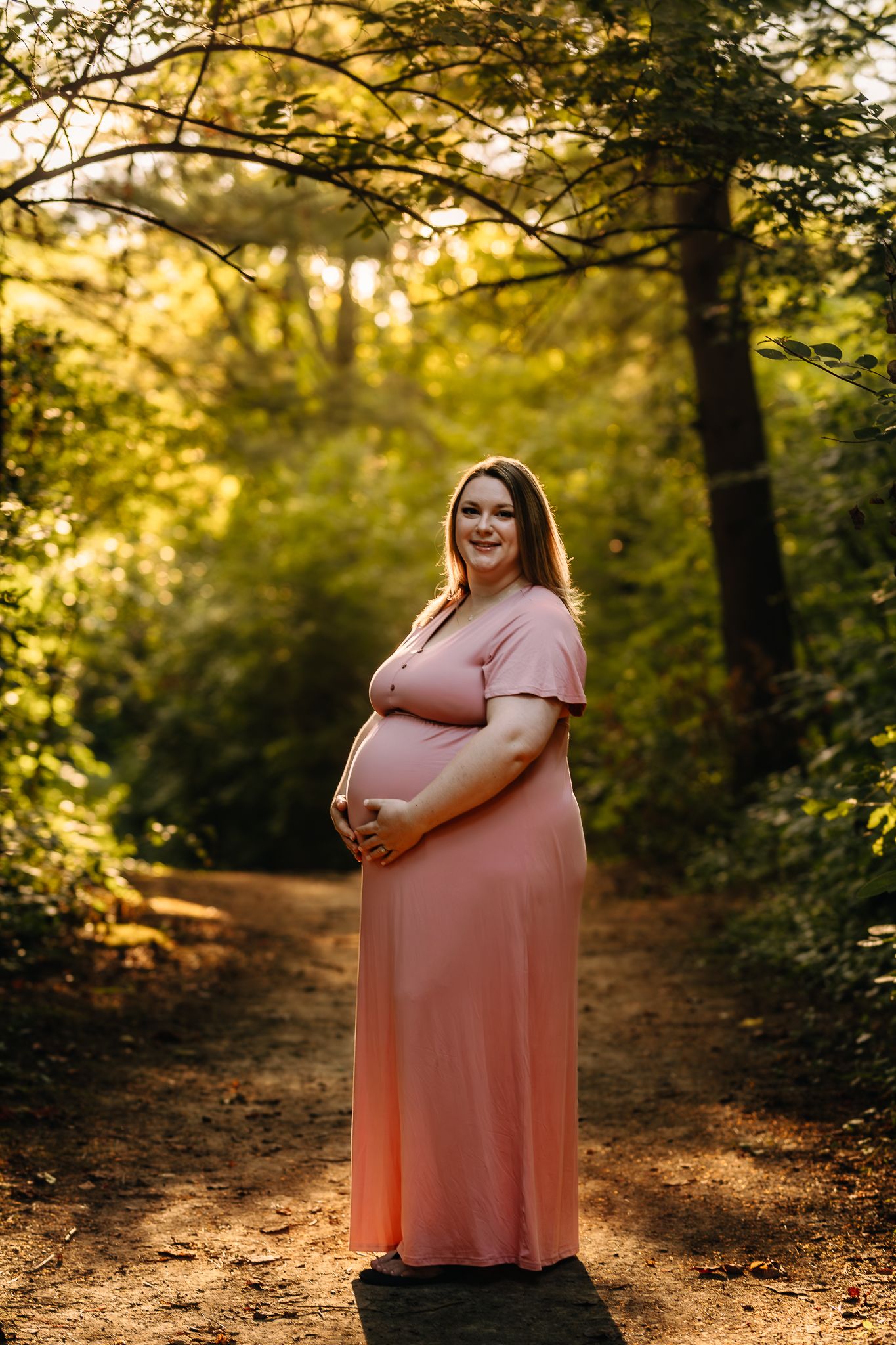 Smiling pregnant woman in pink maxi dress holding baby bump on a sunlit forest path.