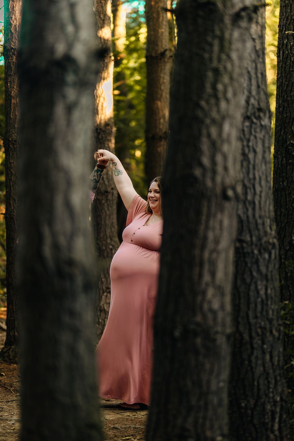 Joyful pregnant woman in pink dress posing among tall trees in a sun-dappled forest.